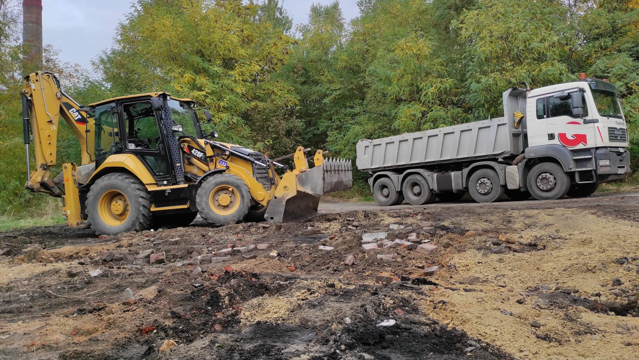 Żółta koparko-ładowarka CAT 428F i biała ciężarówka MAN na placu budowy z gruzem ceglanym i ziemią, w tle drzewa.