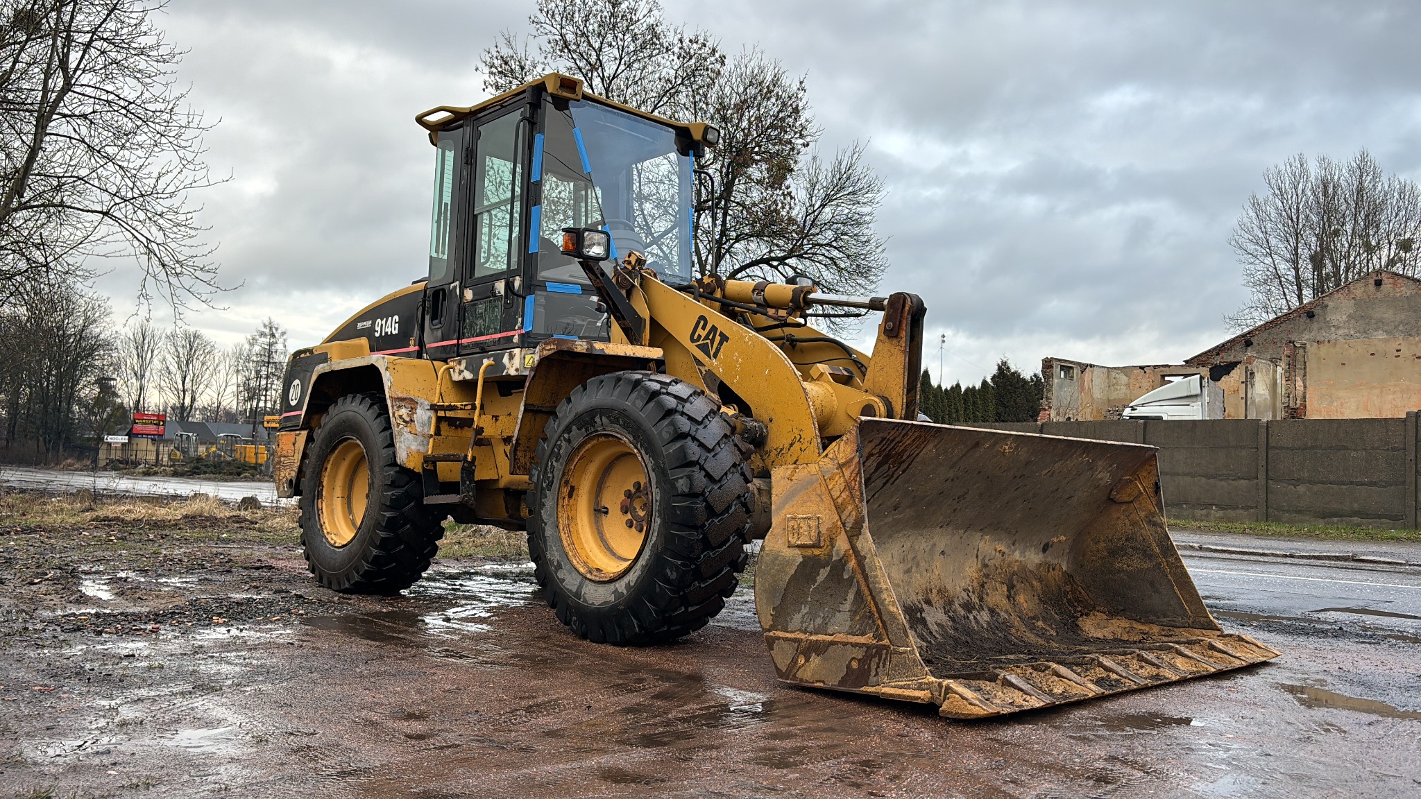 Żółta ładowarka CAT 914G na mokrym asfalcie przed zrujnowanym budynkiem i betonowym płotem, pochmurne niebo w tle.