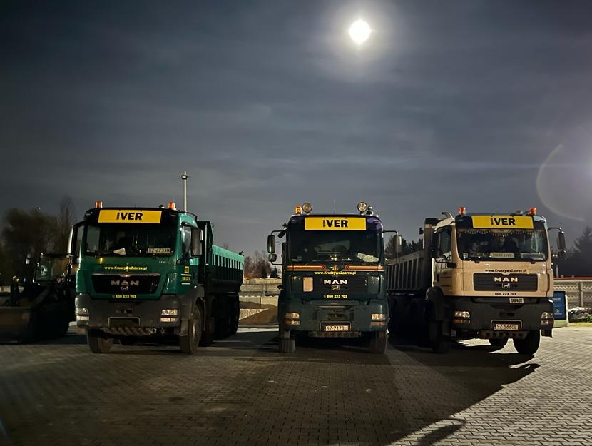 Trzy ciężarówki MAN typu wywrotka, w kolorach zielonym, ciemnozielonym i beżowym, stoją zaparkowane nocą pod jasnym oświetleniem, z widocznym księżycem w tle.
