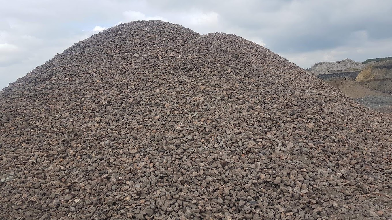 Duży pryzmat szarego kruszywa kamiennego na tle pochmurnego nieba i wyrobiska górniczego w oddali.