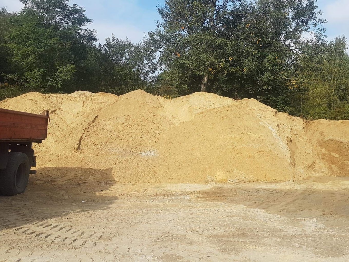 Kopalnia piasku: wysoka pryzma jasnego piasku z widocznymi śladami opon ciężarówki, w tle drzewa liściaste i fragment skrzyni ładunkowej samochodu ciężarowego.