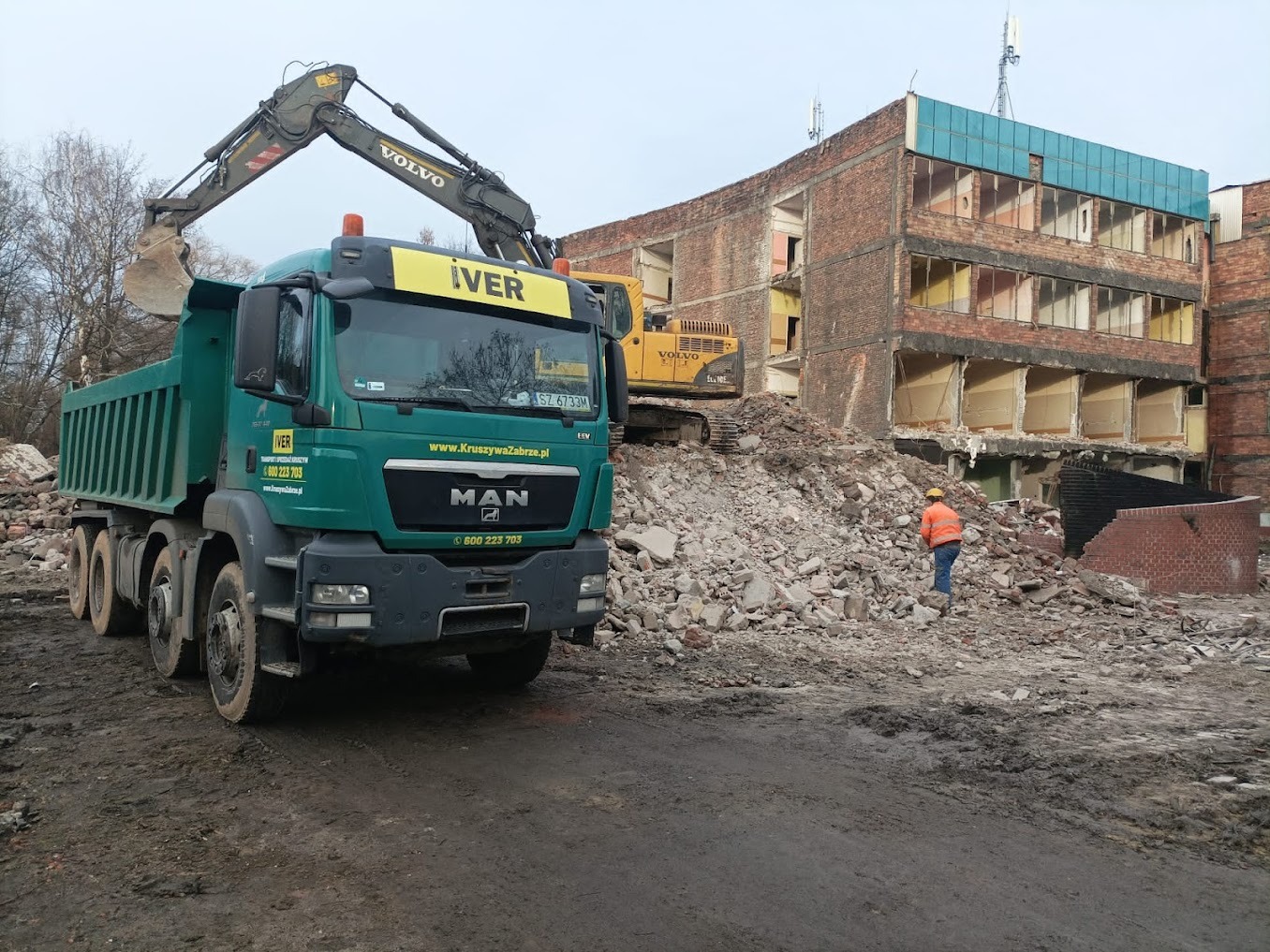 Zielona ciężarówka MAN z koparką Volvo IVER załadowuje gruz z rozbiórki budynku, w tle widoczna żółta koparka i pracownik w pomarańczowej kamizelce.