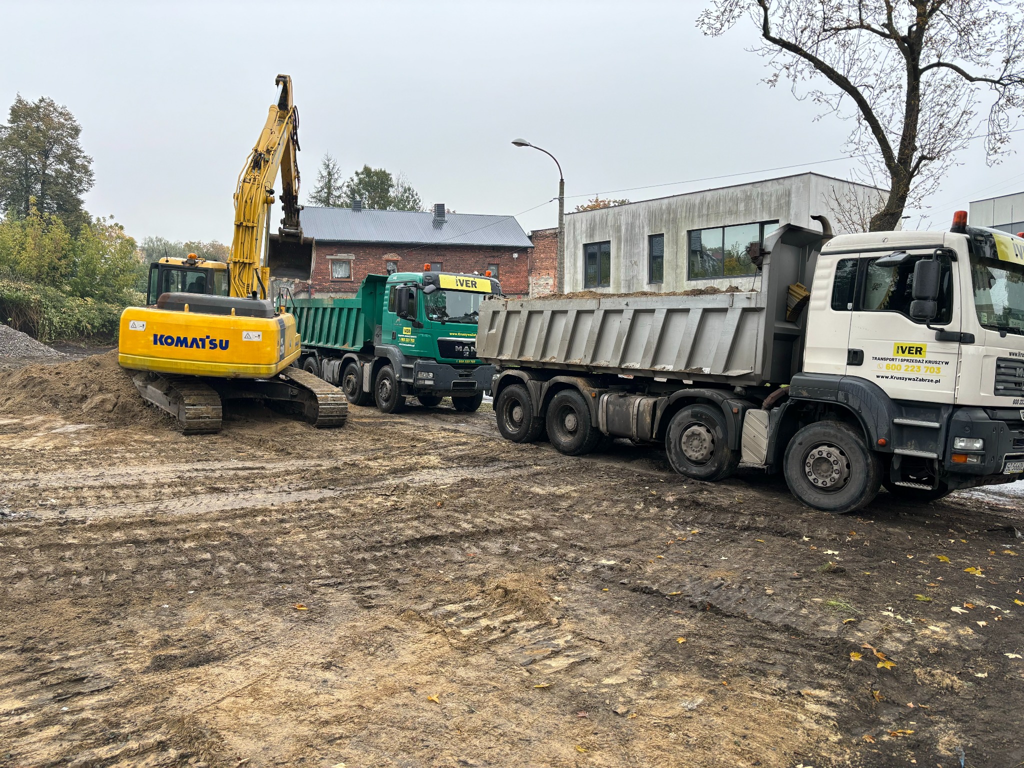 Żółta koparka Komatsu ładuje urobek do dwóch ciężarówek MAN IVER na placu budowy w pochmurny dzień. Widoczne ślady opon w błotnistej ziemi i budynki w tle.