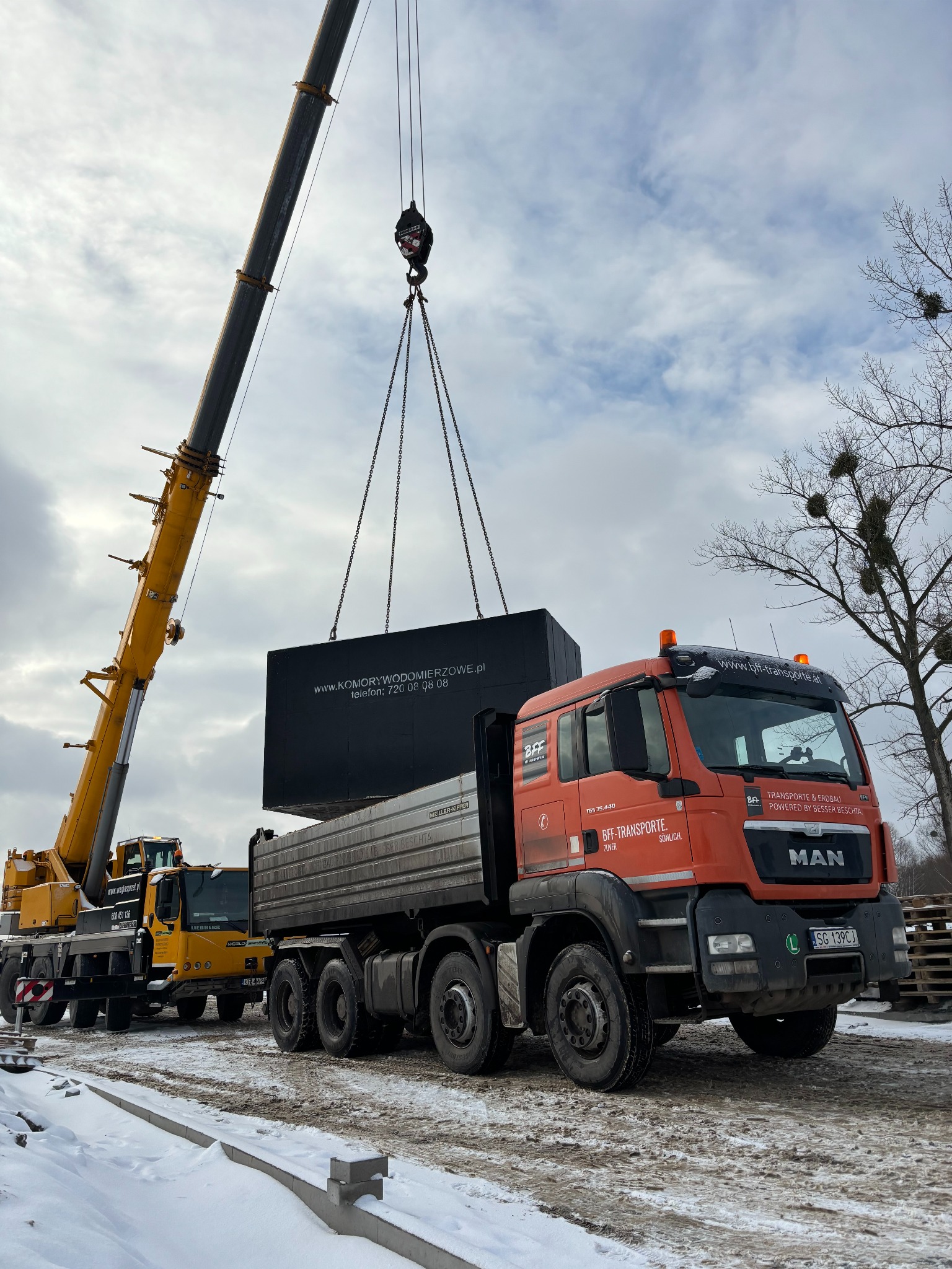 Żuraw przenosi duży, czarny zbiornik na pomarańczową ciężarówkę MAN na placu budowy w zimowej scenerii, w tle widoczne drzewa i pochmurne niebo.