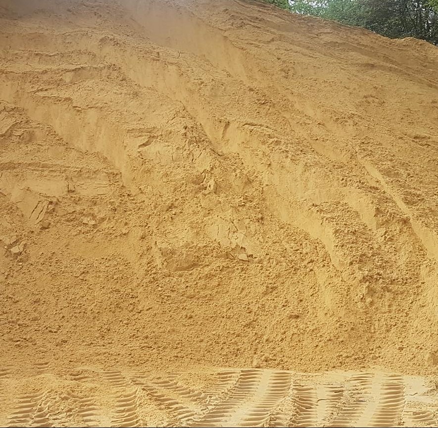 Góra jasnego piasku z wyraźnymi śladami opon pojazdu budowlanego u podstawy, częściowo zacieniona u góry, z fragmentem zieleni w tle.