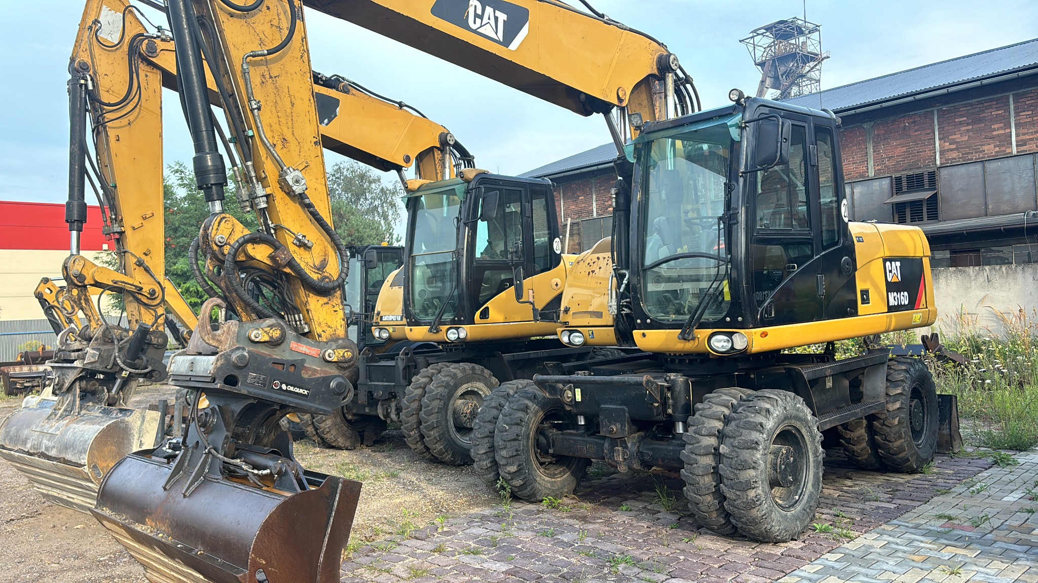 Żółte koparki kołowe marki CAT M316D zaparkowane na brukowanej powierzchni, w tle budynek z cegły i wieża szybu kopalnianego.