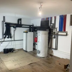 Instalacja pompy ciepła w kotłowni: biała jednostka wewnętrzna, owinięty folią zbiornik buforowy, rury w izolacji termicznej, zabezpieczone podłoże, w tle ściany z instalacją hydrauliczną.