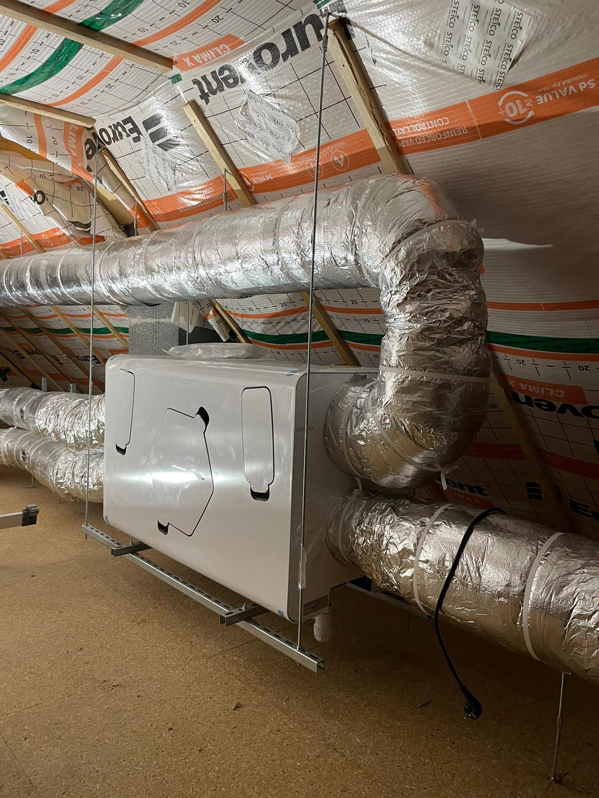 Centrala wentylacyjna z rekuperacją, zamontowana na poddaszu, z widocznymi przewodami wentylacyjnymi owiniętymi folią aluminiową, na tle membrany dachowej z nadrukiem.