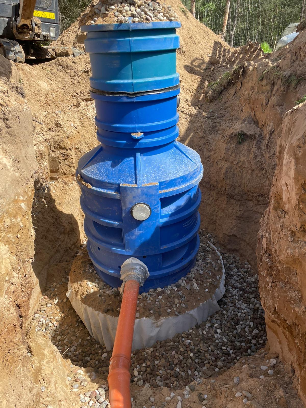 Niebieski zbiornik z tworzywa sztucznego w wykopie, obsypany żwirem, z podłączoną pomarańczową rurą, widoczny fragment koparki w tle.