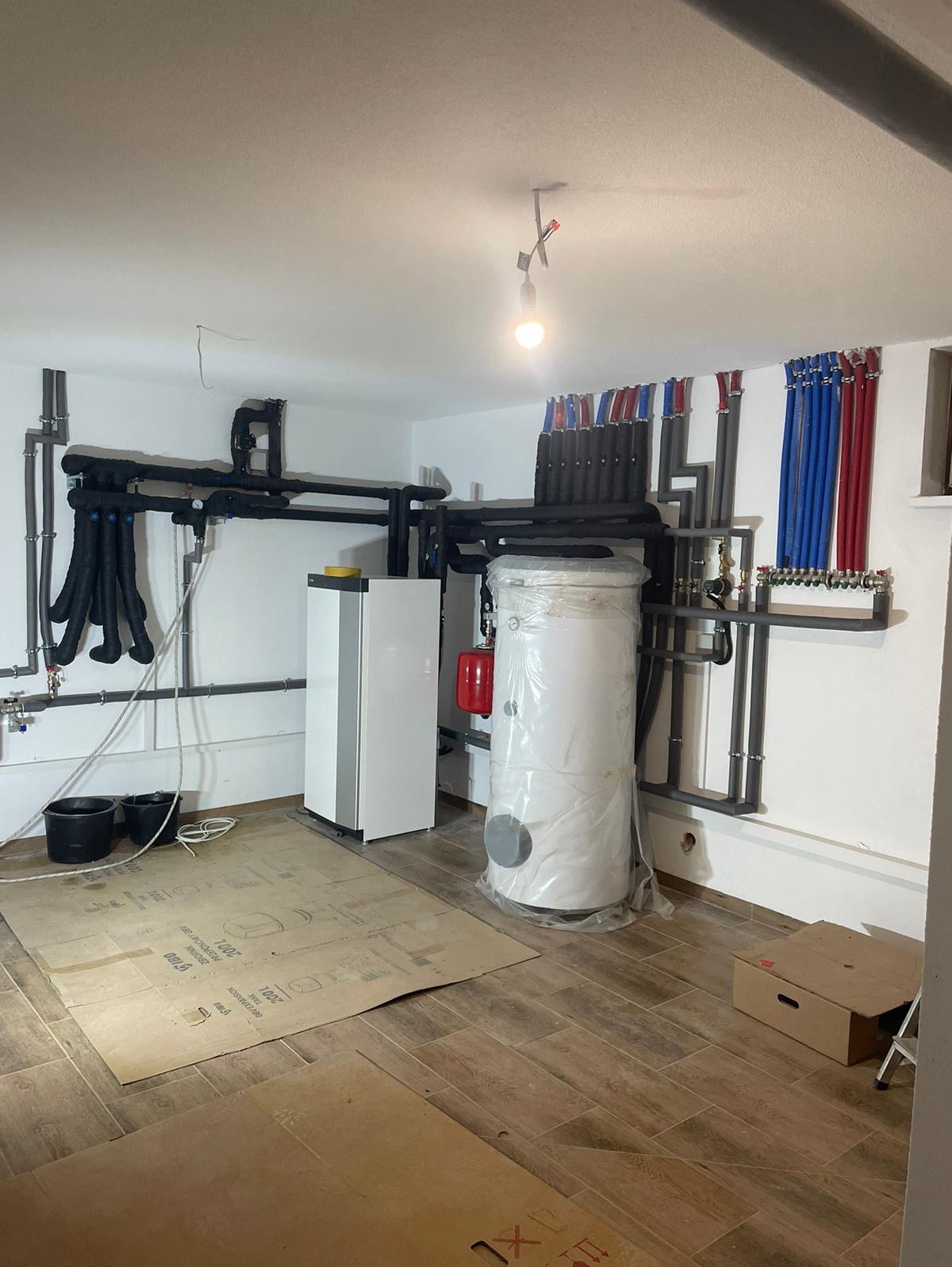 Instalacja pompy ciepła w kotłowni: biała jednostka wewnętrzna, owinięty folią zbiornik buforowy, rury w izolacji termicznej, zabezpieczone podłoże, w tle ściany z instalacją hydrauliczną.