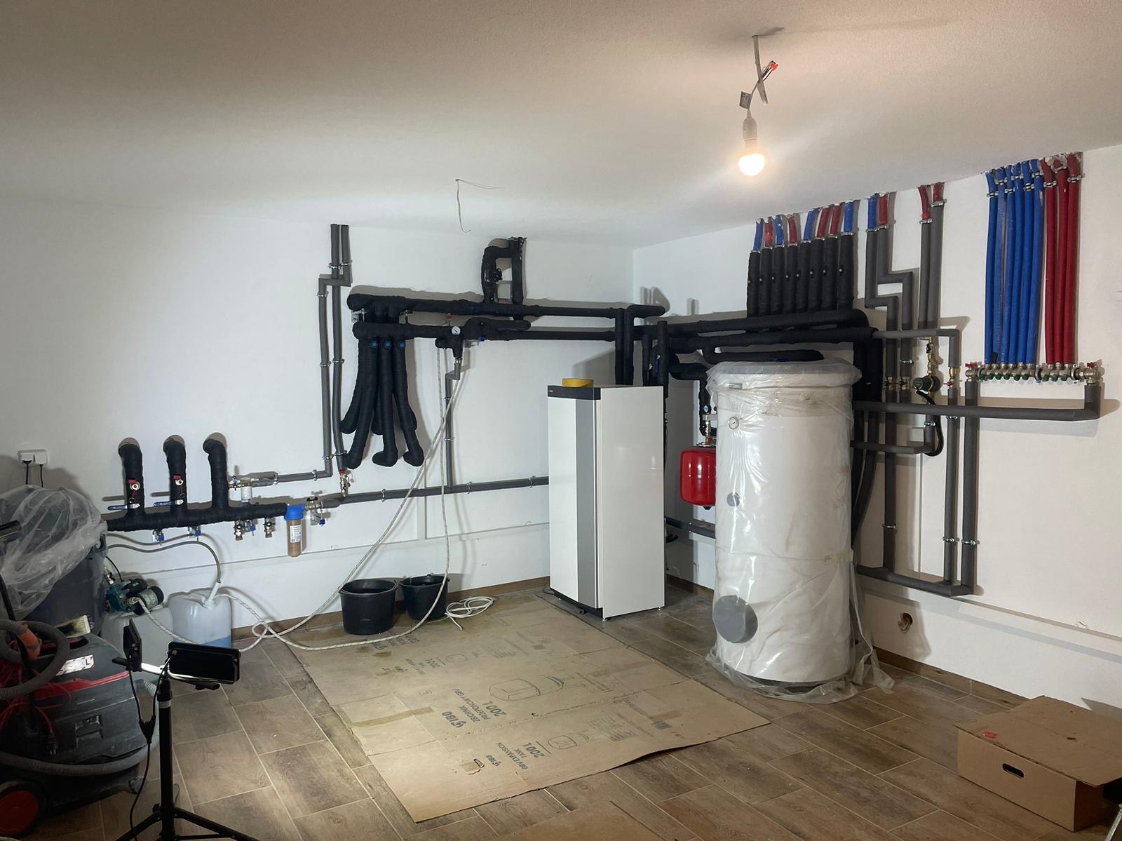 Wnętrze kotłowni z zainstalowanym nowoczesnym piecem grzewczym, dużym zbiornikiem na wodę owiniętym folią ochronną, rozbudowaną instalacją rur w izolacji termicznej oraz elementami osprzętu...