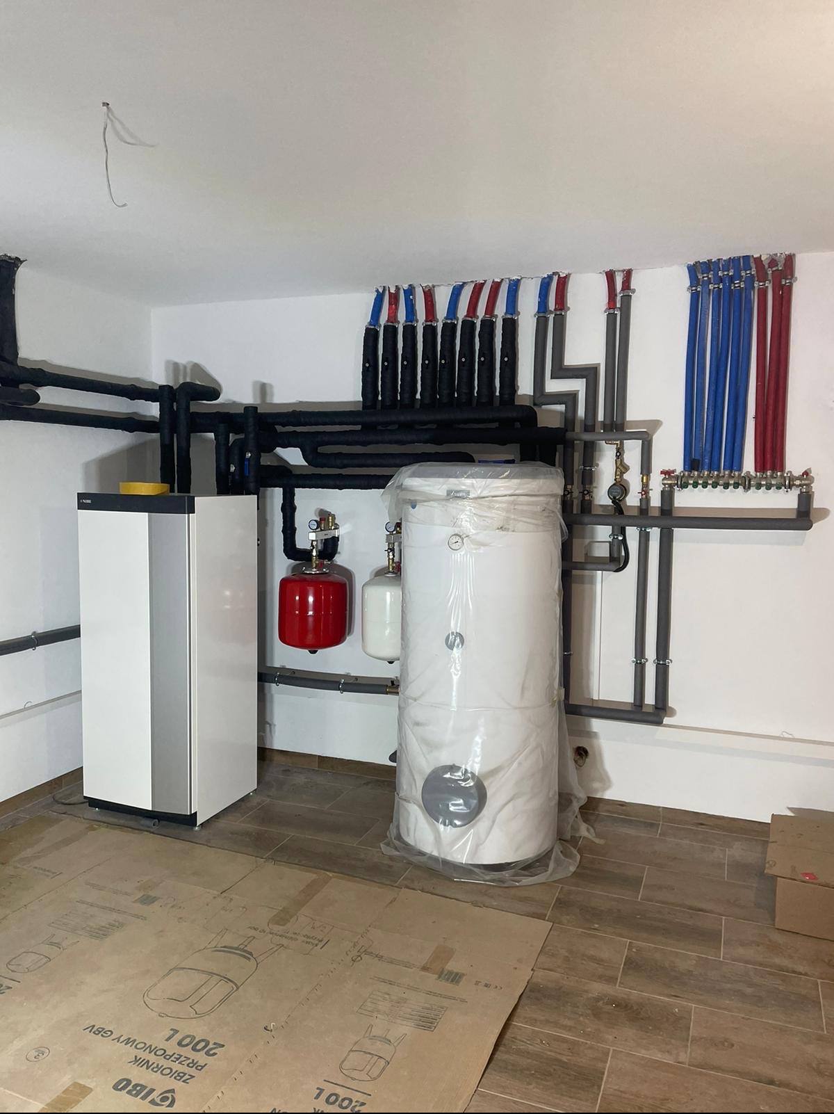 Zainstalowany system grzewczy z kotłem, zbiornikiem buforowym w folii i rurami w izolacji, widoczne także małe zbiorniki wyrównawcze w kolorze czerwonym i białym.