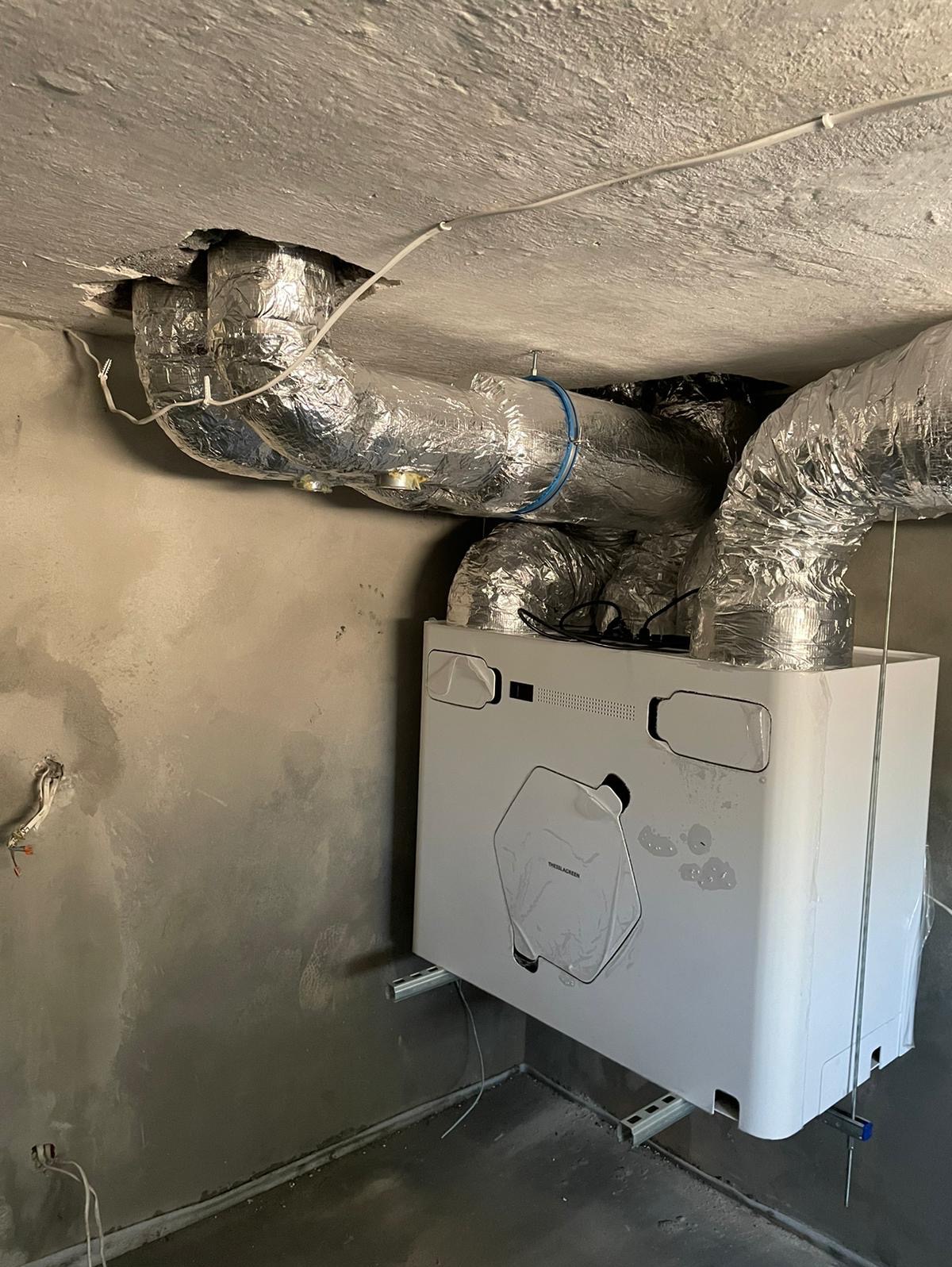 Biała jednostka rekuperacyjna zamontowana na ścianie w surowym wnętrzu, z widocznymi przewodami wentylacyjnymi owiniętymi folią aluminiową oraz elementami instalacji elektrycznej.