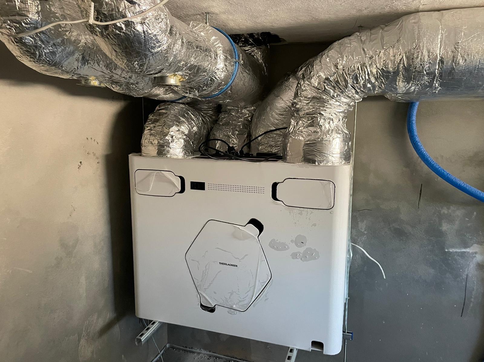 Centrala wentylacyjna z rekuperacją, podłączona do aluminiowych przewodów wentylacyjnych, widoczna na tle surowej ściany w trakcie montażu.