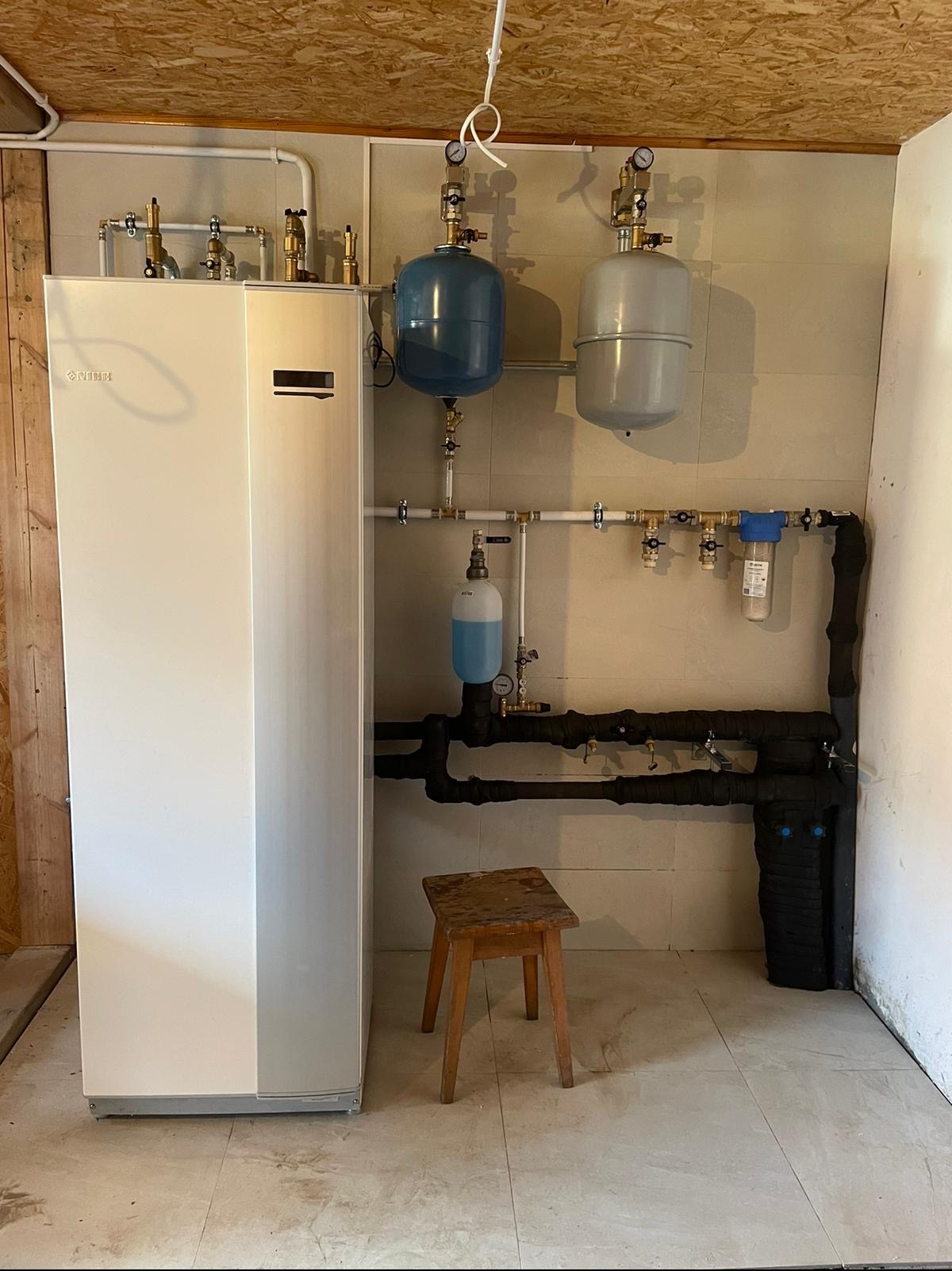 Biała jednostka grzewcza NIBE z dwoma zbiornikami wyrównawczymi (niebieski i szary) i rurami w izolacji, na tle wykafelkowanej ściany i surowego drewnianego sufitu, obok stoi drewniany stołek.