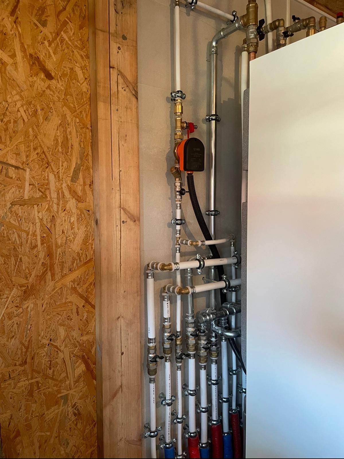 Złożony system rur instalacji grzewczej z pompą obiegową zamontowany na ścianie z płyt OSB, widoczne liczne zawory i złączki.