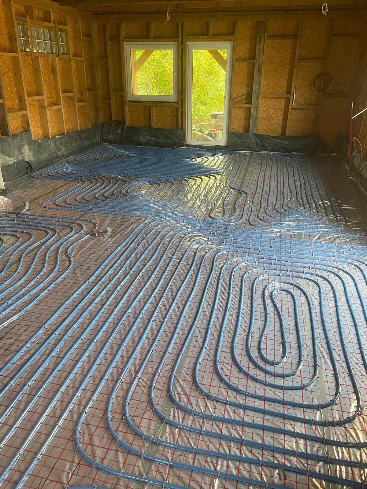 Montaż niebieskich rur ogrzewania podłogowego na folii z siatką w surowym wnętrzu budynku w Białystoku, widoczne okna i drewniana konstrukcja ścian.