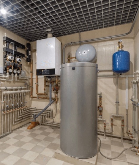 Kompaktowa kotłownia z białym kotłem gazowym, dużym srebrnym zasobnikiem ciepłej wody i niebieskim zbiornikiem wyrównawczym, widoczne liczne rury miedziane i zawory, całość w pomieszczeniu...