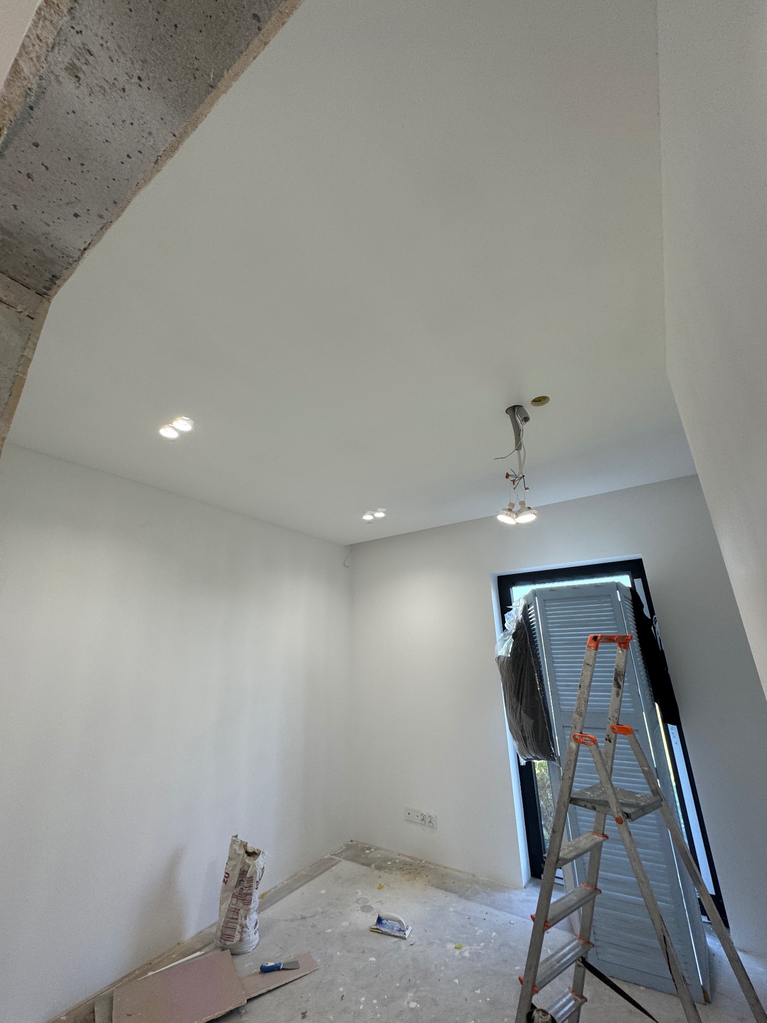 Wnętrze w trakcie remontu: surowy betonowy łuk, świeżo pomalowane białe ściany, zamontowane halogeny w suficie, drabina oparta o okno z żaluzjami, worek gładzi na podłodze.