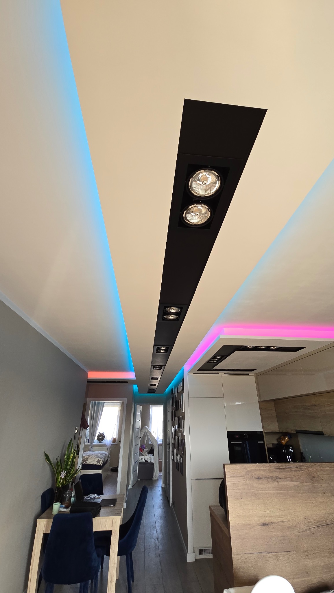 Nowoczesne oświetlenie sufitowe z reflektorami w czarnych obudowach i dekoracyjnym podświetleniem LED w kolorach niebieskim i różowym, widoczne z perspektywy wnętrza mieszkania.