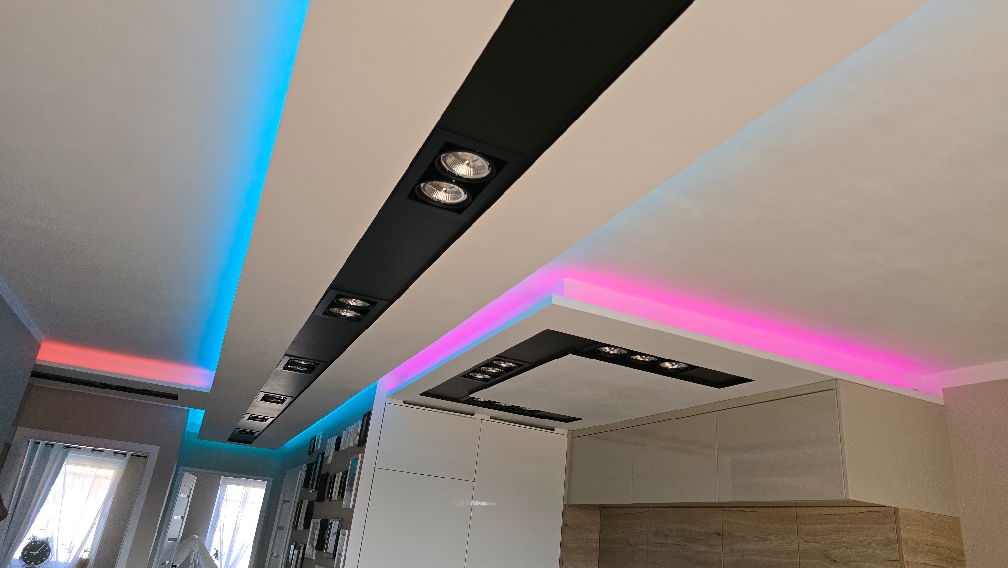 Sufit z podwieszanymi panelami z czarnymi wnękami i punktowym oświetleniem, dodatkowo podświetlony listwami LED w kolorach niebieskim i różowym, widok z dołu.
