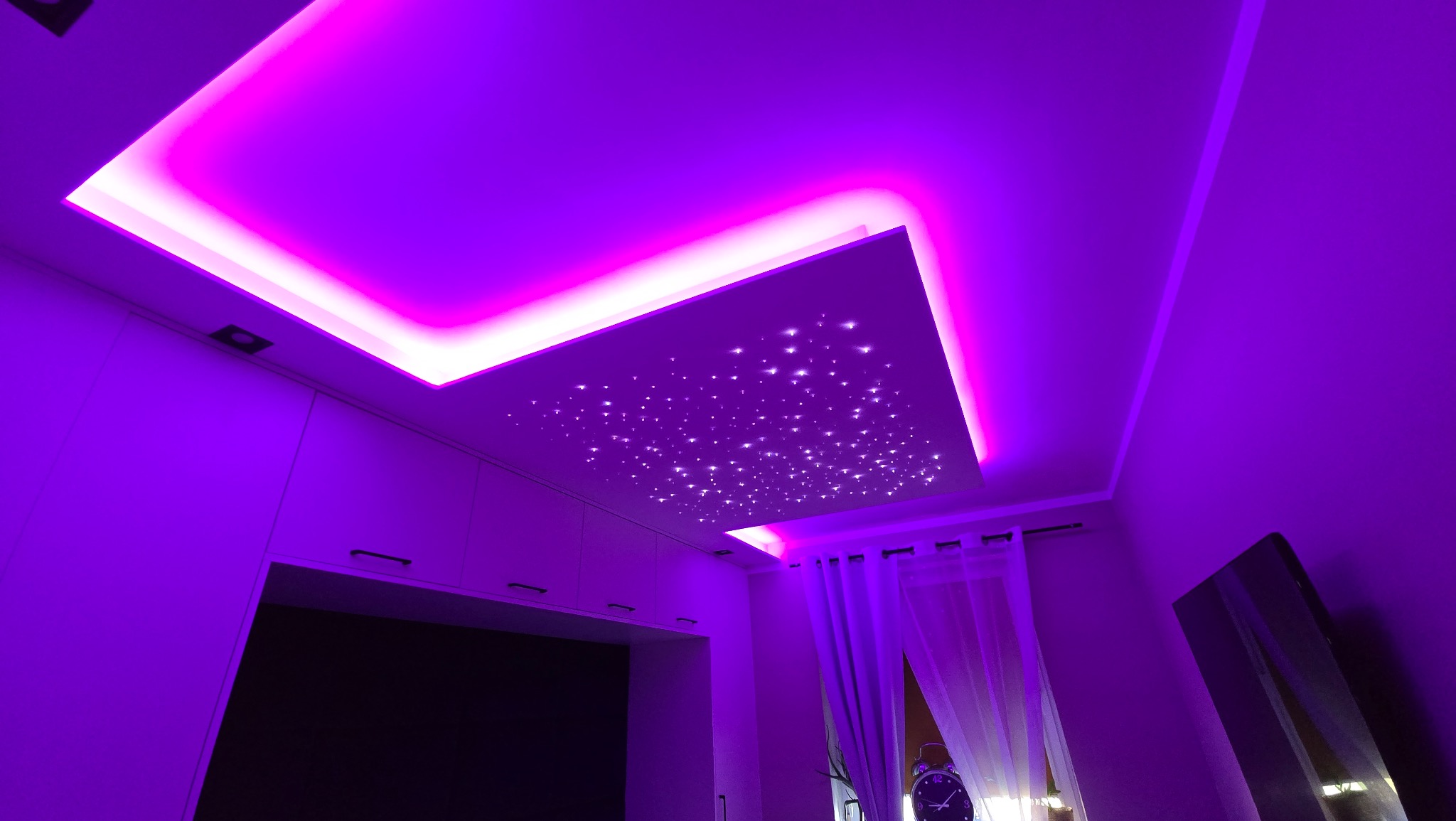 Sufit podwieszany z oświetleniem LED RGB i dekoracyjnymi gwiazdami, w fioletowej tonacji, widoczne meble w tle.