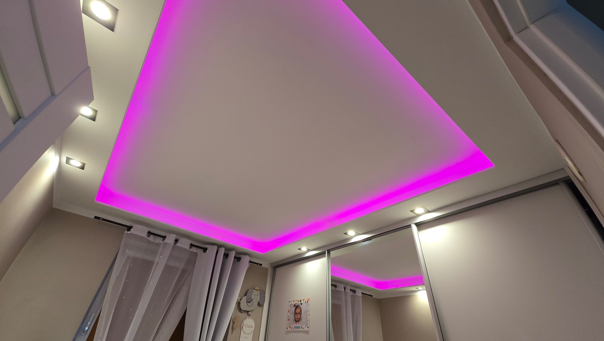 Podwieszany sufit z oświetleniem LED w kolorze fuksji, widok od dołu, w tle zasłony i szafa z lustrem.