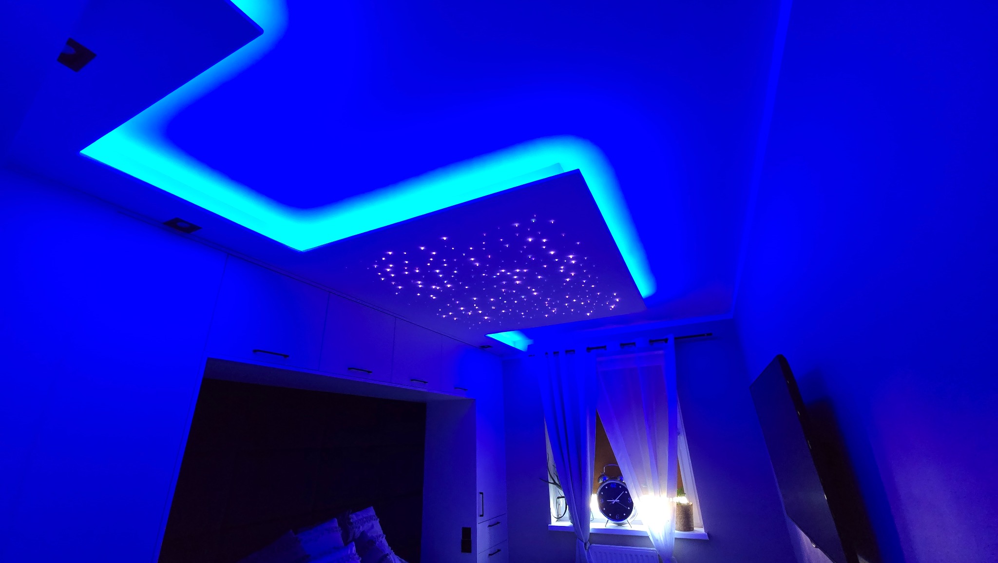 Sufit podwieszany z oświetleniem LED w kolorze niebieskim i imitacją gwiazd, widoczny fragment szafy wnękowej i okno z zasłonami.