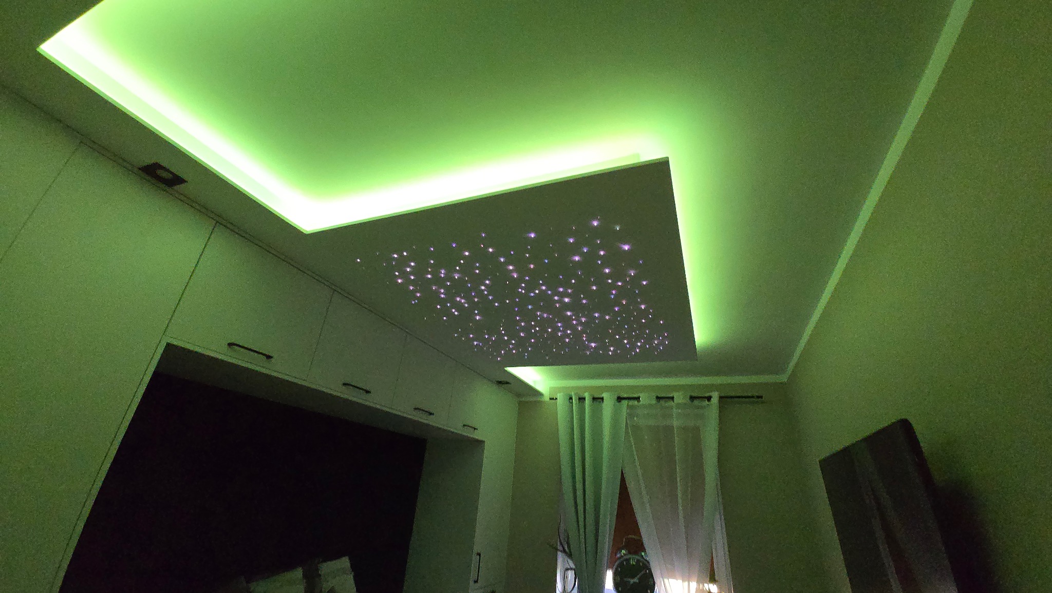 Sufit z dekoracyjnym oświetleniem LED w kolorze zielonym, imitującym gwieździste niebo, w pokoju z zabudową meblową i zasłonami.