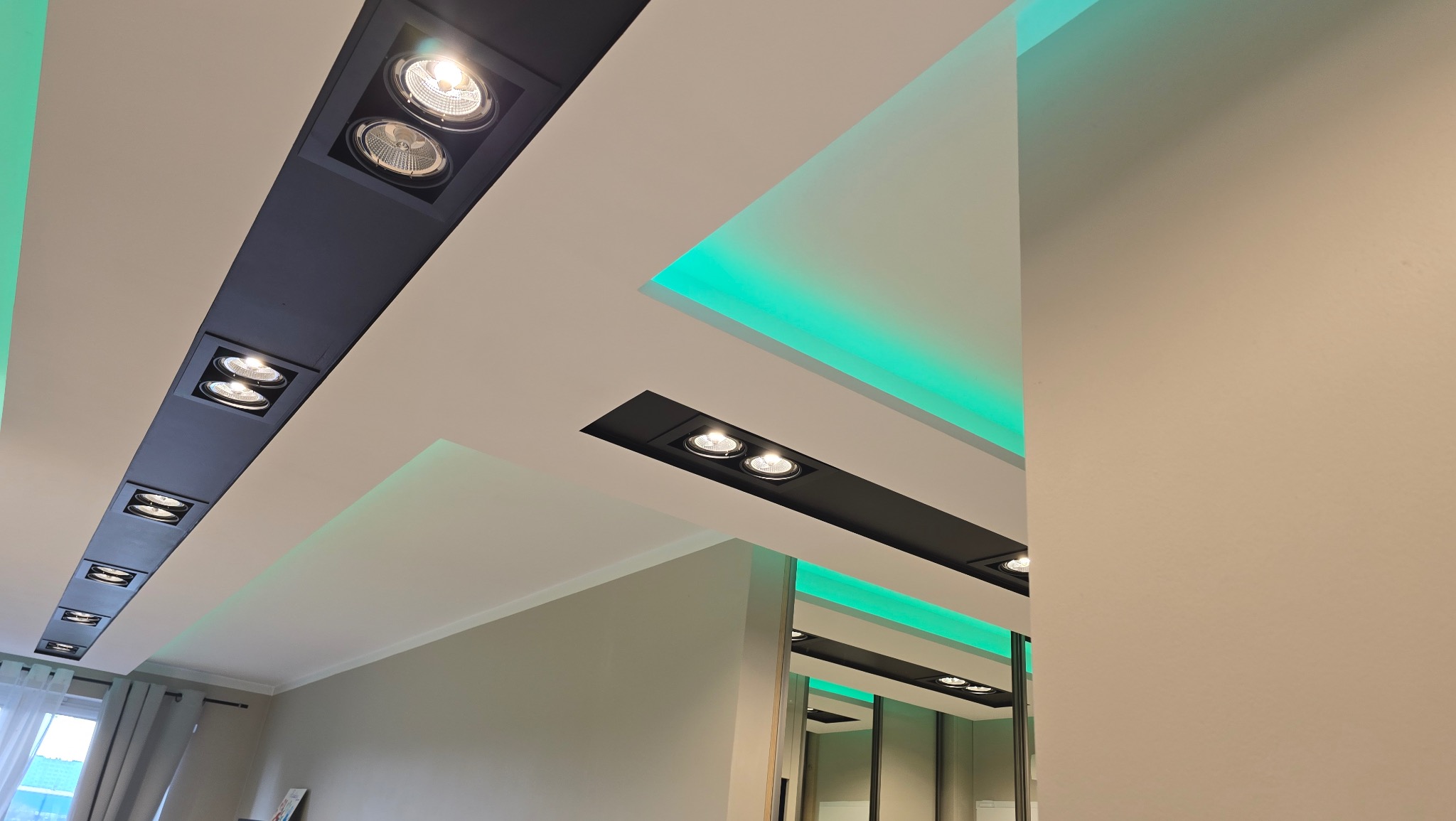 Nowoczesny sufit z wpuszczonymi, czarnymi listwami oświetleniowymi z białymi reflektorami punktowymi i zielonym oświetleniem LED, widok z dołu.