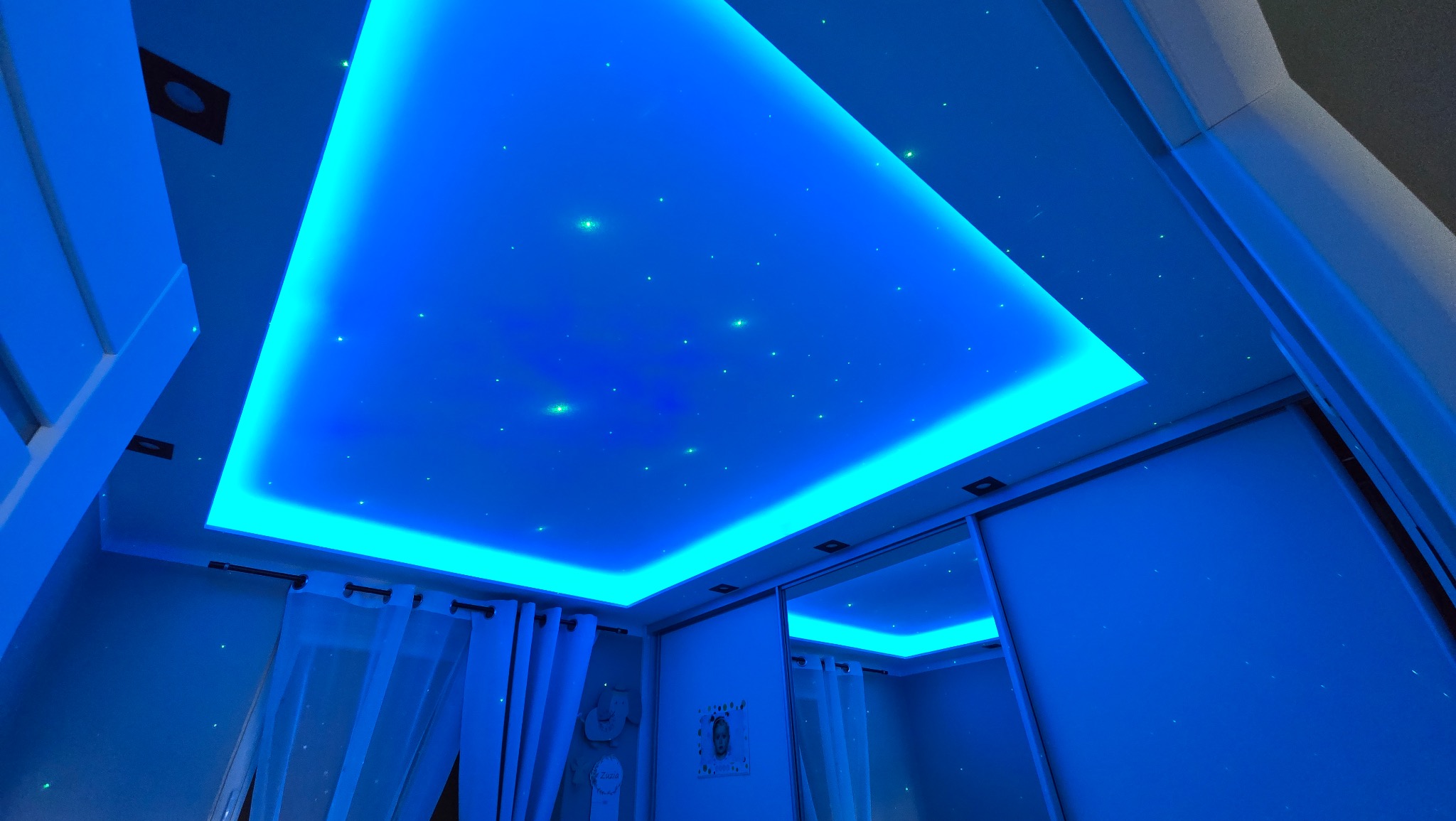 Sufit podwieszany z efektem gwieździstego nieba, oświetlony intensywnym, niebieskim światłem LED, widoczny fragment okna z firanką i szafa przesuwna z lustrem.