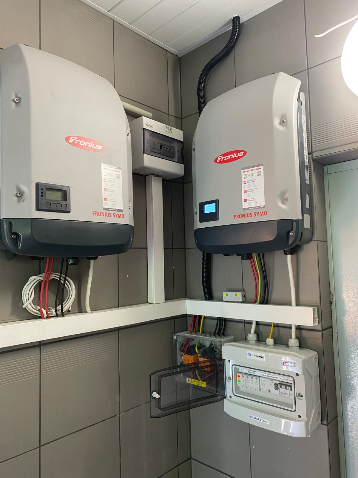 Dwa falowniki Fronius Symo zamontowane na ścianie z szarych płytek, widoczne kable i rozdzielnica AC z otwartą osłoną, instalacja fotowoltaiczna w pomieszczeniu.