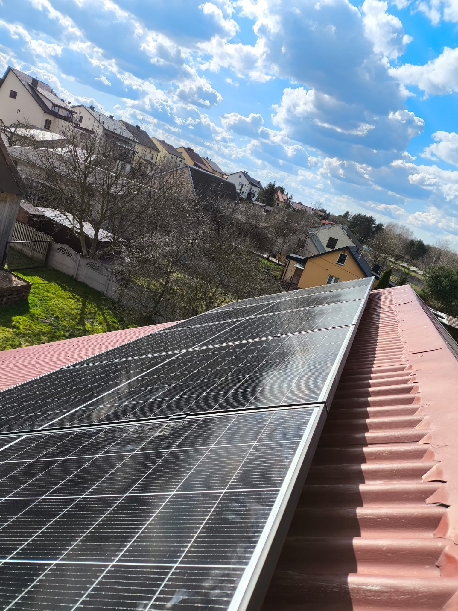Panele słoneczne zamontowane na falistej, pomalowanej na czerwono blasze dachowej, w tle zabudowa jednorodzinna i błękitne niebo z chmurami.