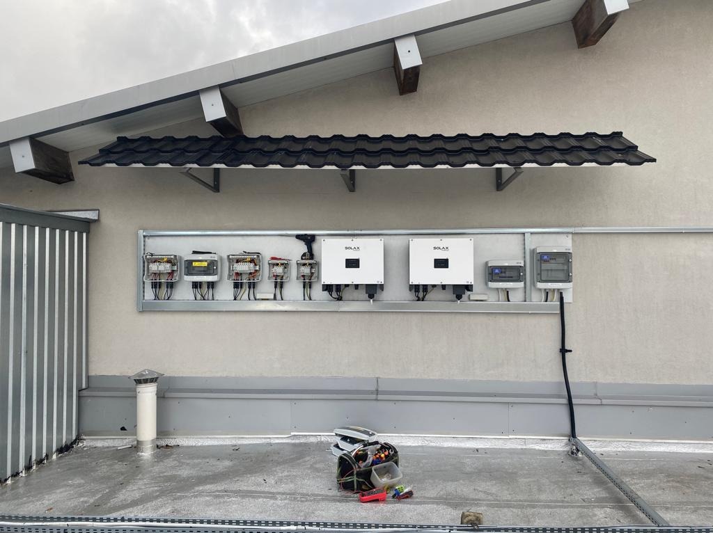 Instalacja fotowoltaiczna na dachu: widoczne inwertery marki Solax, skrzynki elektryczne w metalowej ramie, narzędzia na dachu, fragment dachu z dachówką ceramiczną.