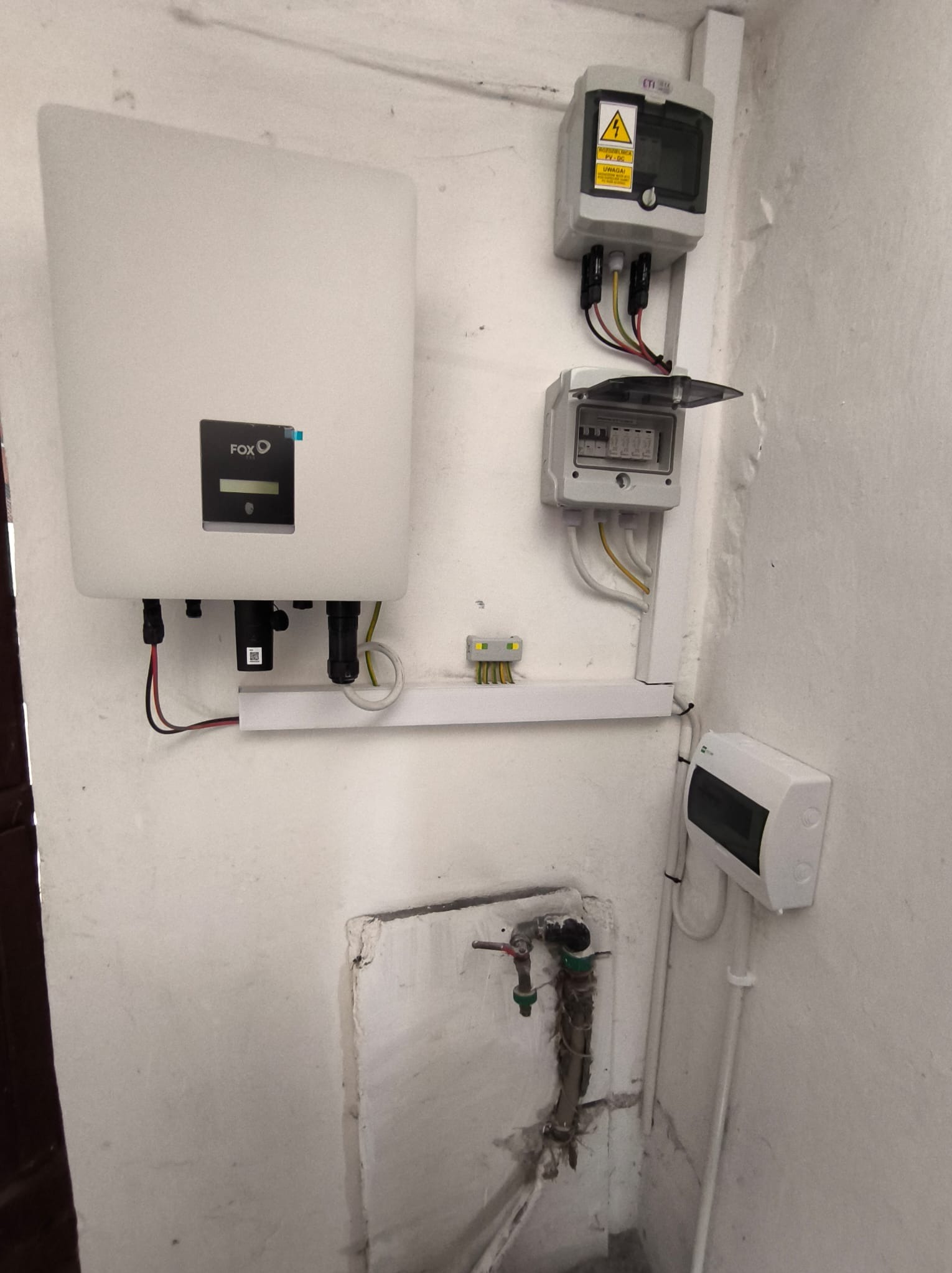 Biały falownik FOX z wyświetlaczem, połączony z instalacją elektryczną wewnątrz budynku, widoczne zabezpieczenia nadprądowe i skrzynka rozdzielcza.