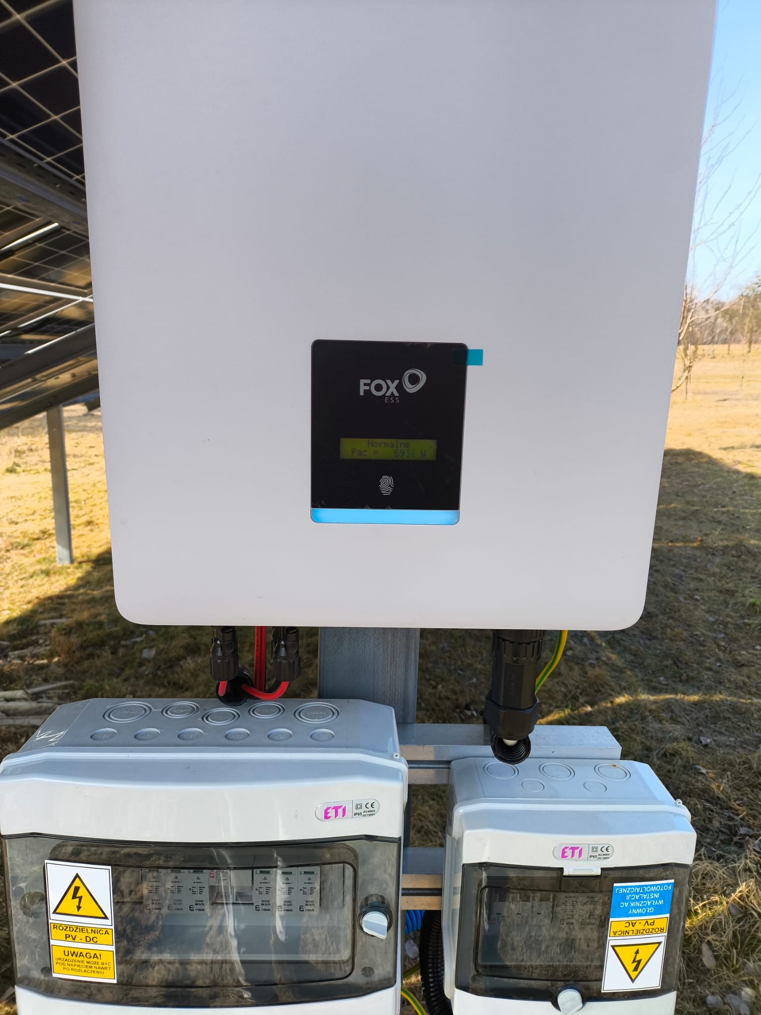 Biały inwerter fotowoltaiczny FoxESS z wyświetlaczem wskazującym bieżącą moc, zamontowany na zewnątrz obok dwóch szarych rozdzielnic ETI z ostrzeżeniami o napięciu, na tle trawy i drzew.