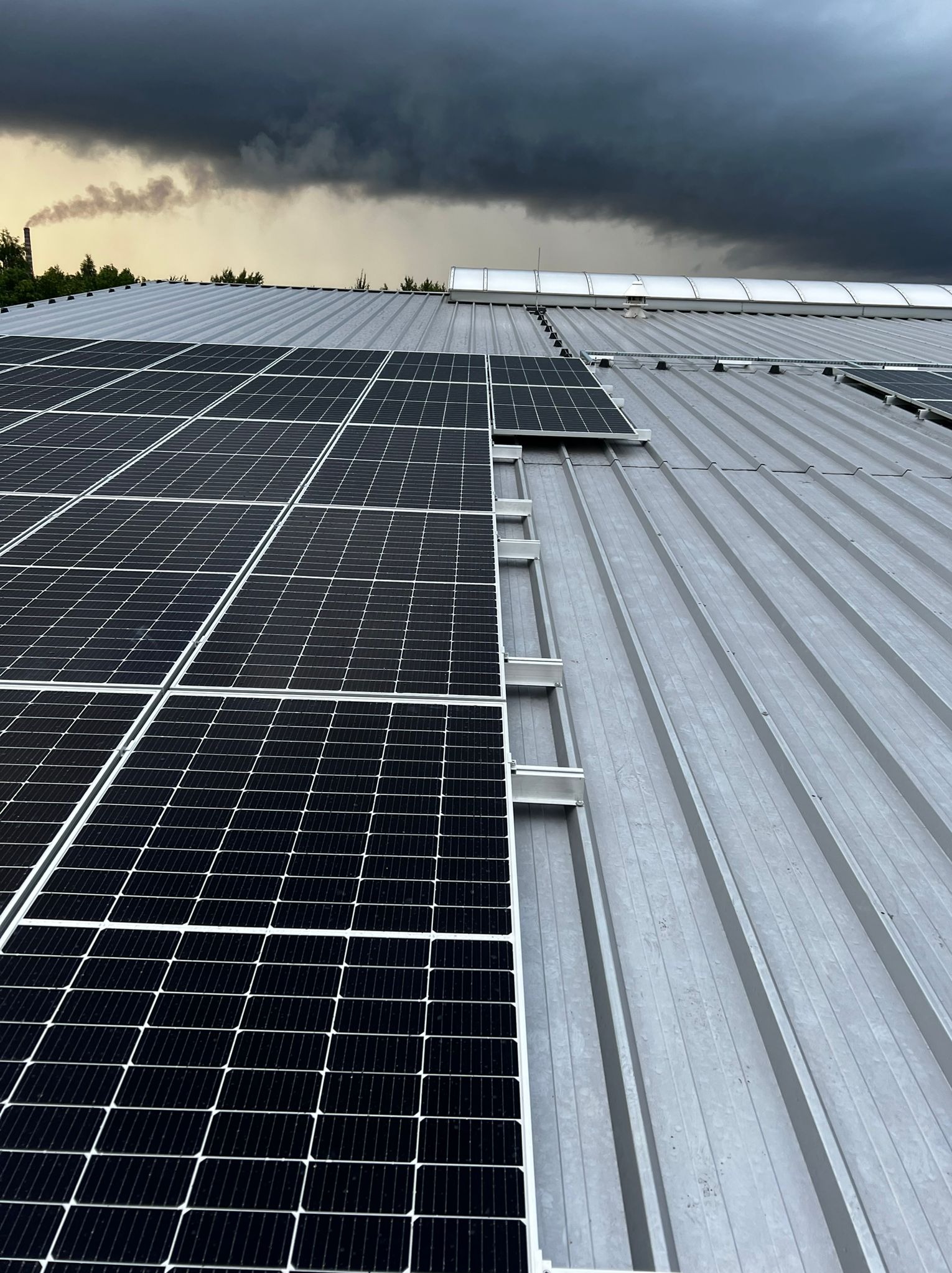 Częściowo zamontowane panele słoneczne na metalowym dachu z widocznym kominem fabrycznym w tle pod pochmurnym niebem.