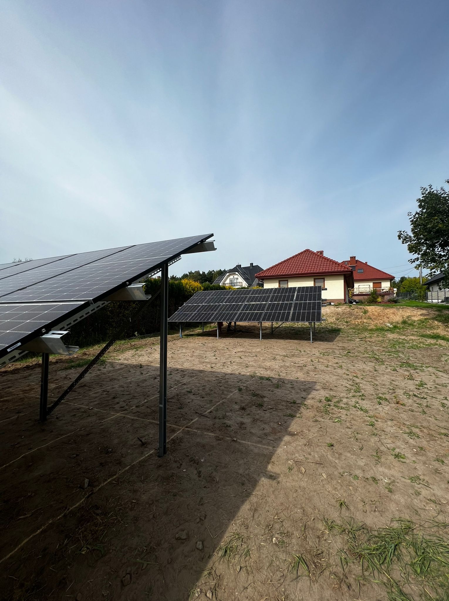 Dwa rzędy paneli słonecznych na metalowych konstrukcjach, widok z ziemi w kierunku domów jednorodzinnych w tle, instalacja w trakcie realizacji.