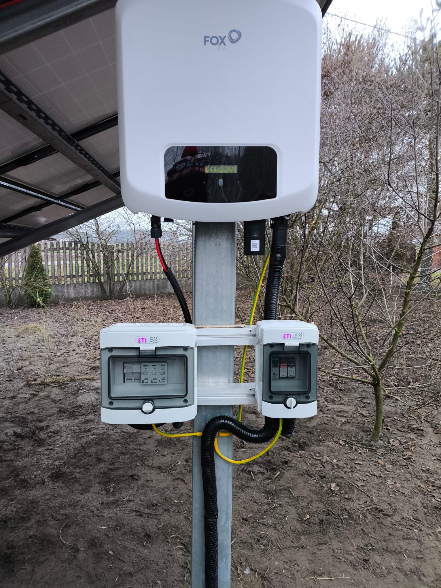 Biały inwerter FoxESS zamontowany na metalowym słupie obok paneli fotowoltaicznych, z dwoma skrzynkami ETI zabezpieczającymi instalację, widoczne przewody ochronne i czarna rura karbowana, tło...