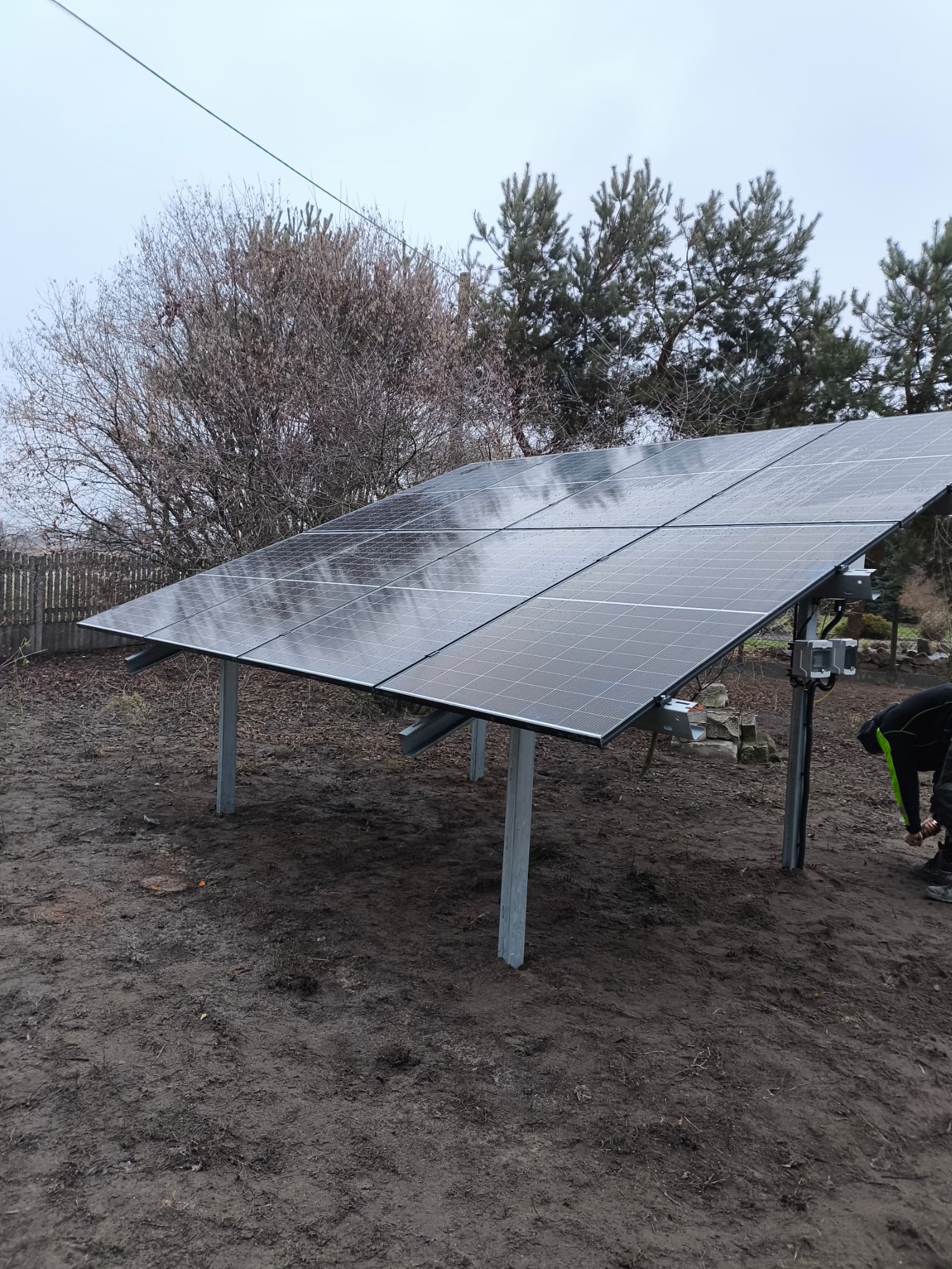 Montaż paneli słonecznych na metalowej konstrukcji, widoczne krople deszczu na panelach, w tle drzewa i drewniany płot, pracownik w trakcie instalacji.