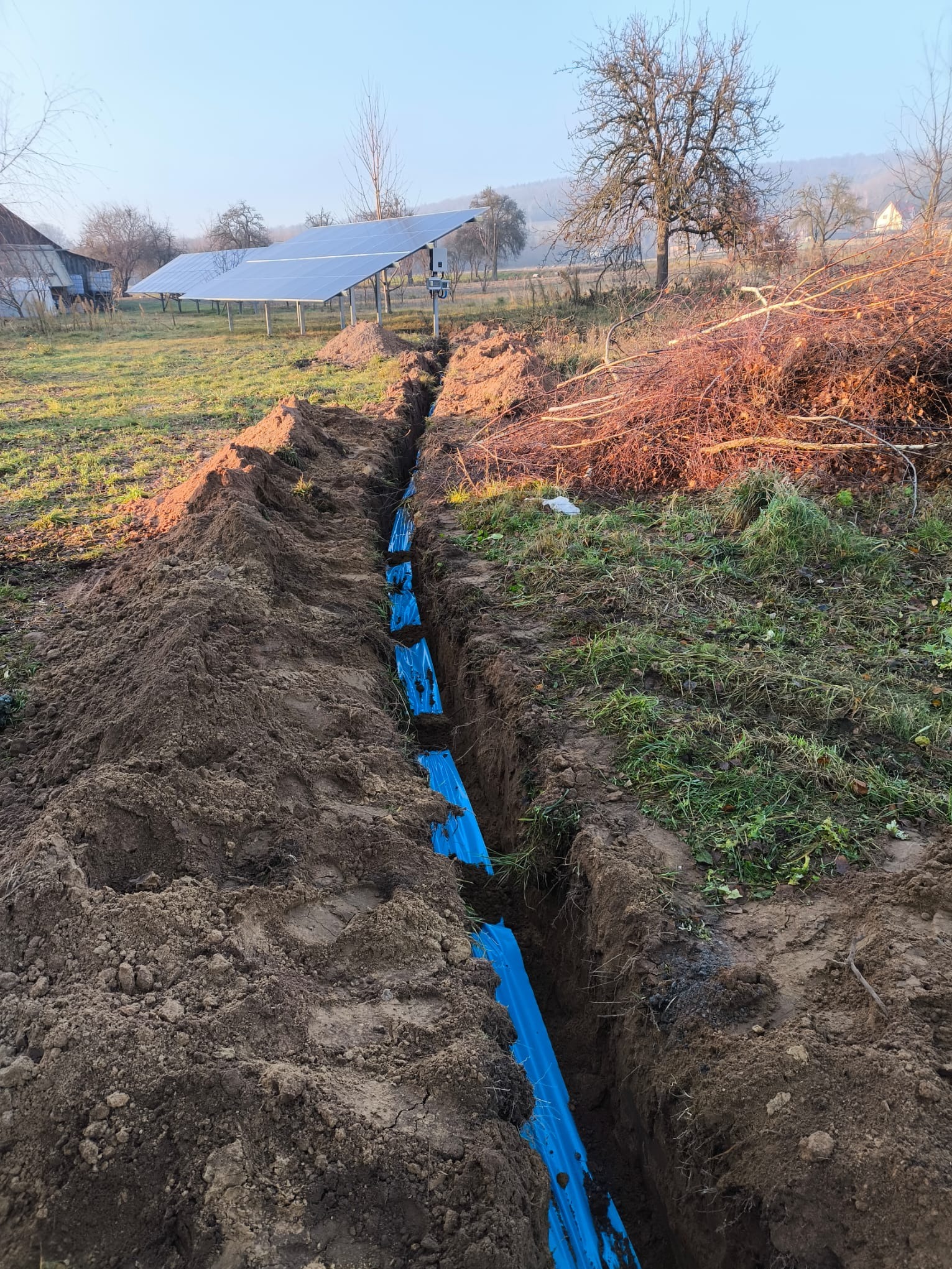 Wykop w ziemi z niebieską rurą osłonową, w tle panele słoneczne i budynek gospodarczy w wiejskim krajobrazie.