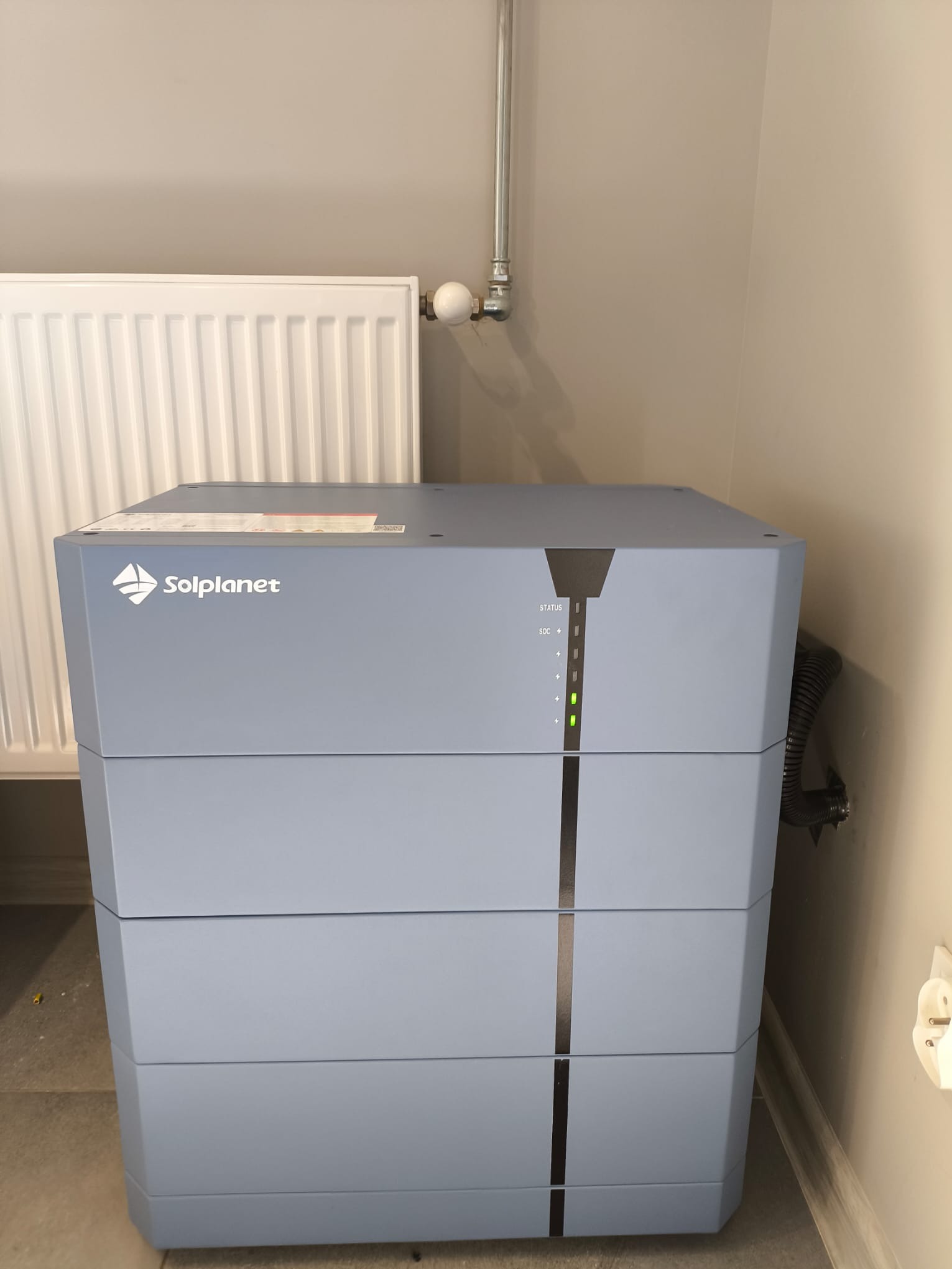 Szary moduł magazynu energii Solplanet, z widocznym logo i diodami statusu, zainstalowany w pomieszczeniu z grzejnikiem i rurami.