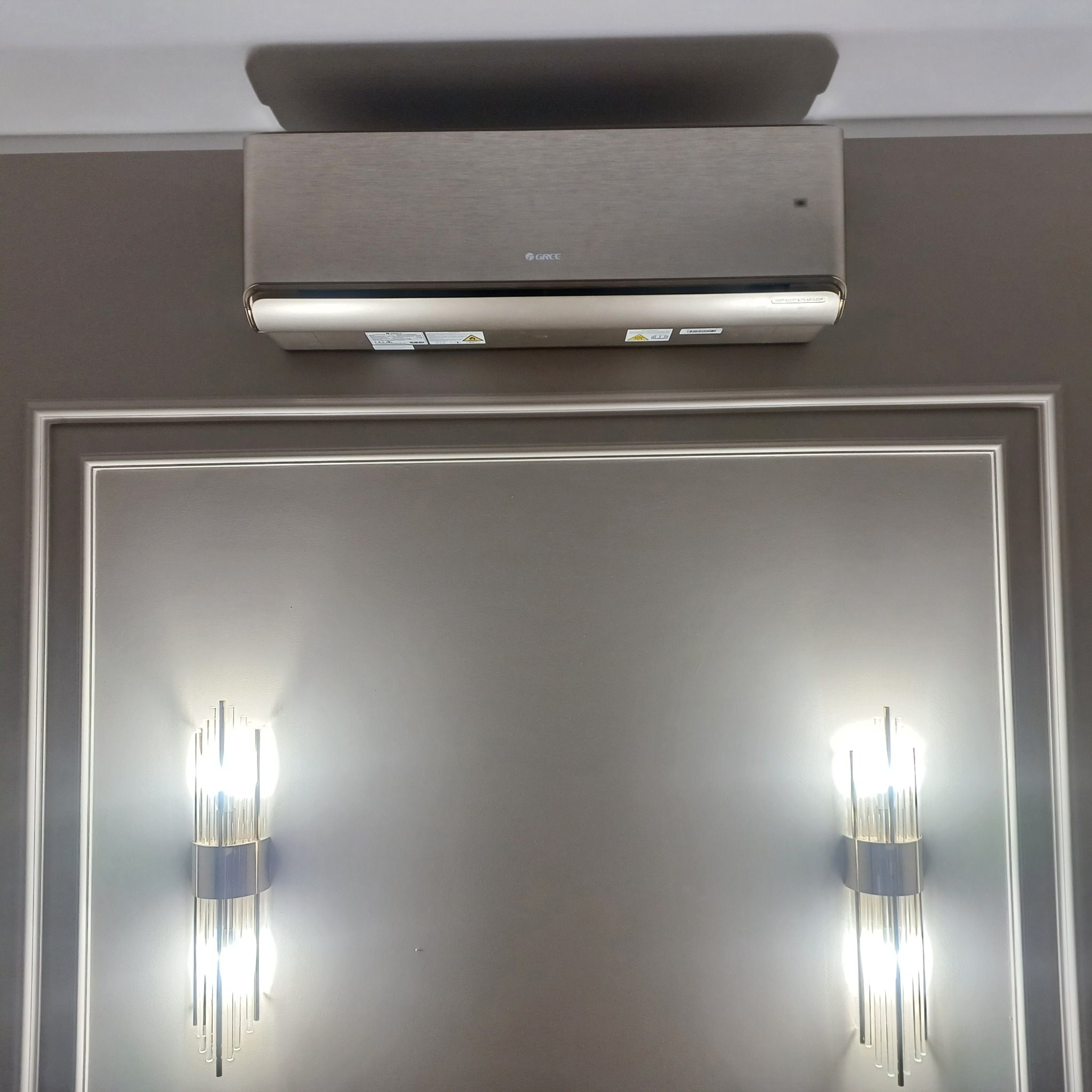 Klimatyzator marki Gree zamontowany na szarej ścianie z dekoracyjną ramą i dwoma szklanymi kinkietami. Widok z przodu, oświetlenie podkreśla detale.