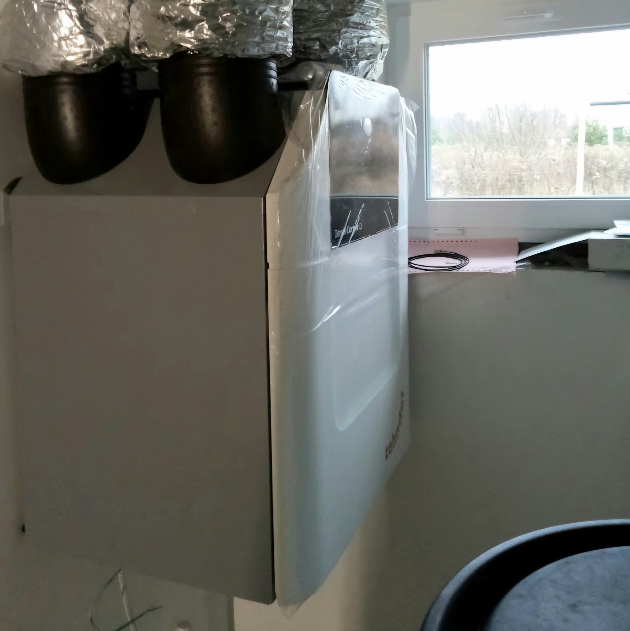 Zamontowany rekuperator Zehnder ComfoAir Q z kanałami wentylacyjnymi w folii ochronnej, widoczne okno i fragment pomieszczenia. Precyzyjny montaż instalacji.