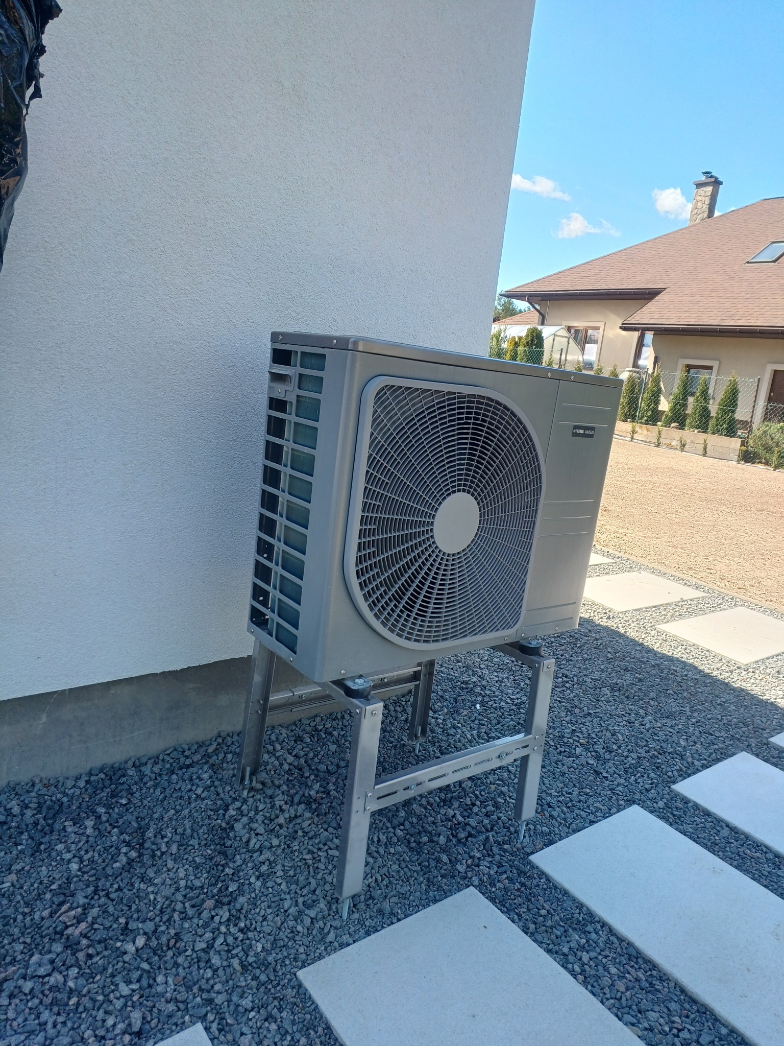 Szara pompa ciepła na metalowym stelażu, zamontowana przy białej ścianie domu. Widoczne szare kamienie i płyty chodnikowe. Błękitne niebo w tle.