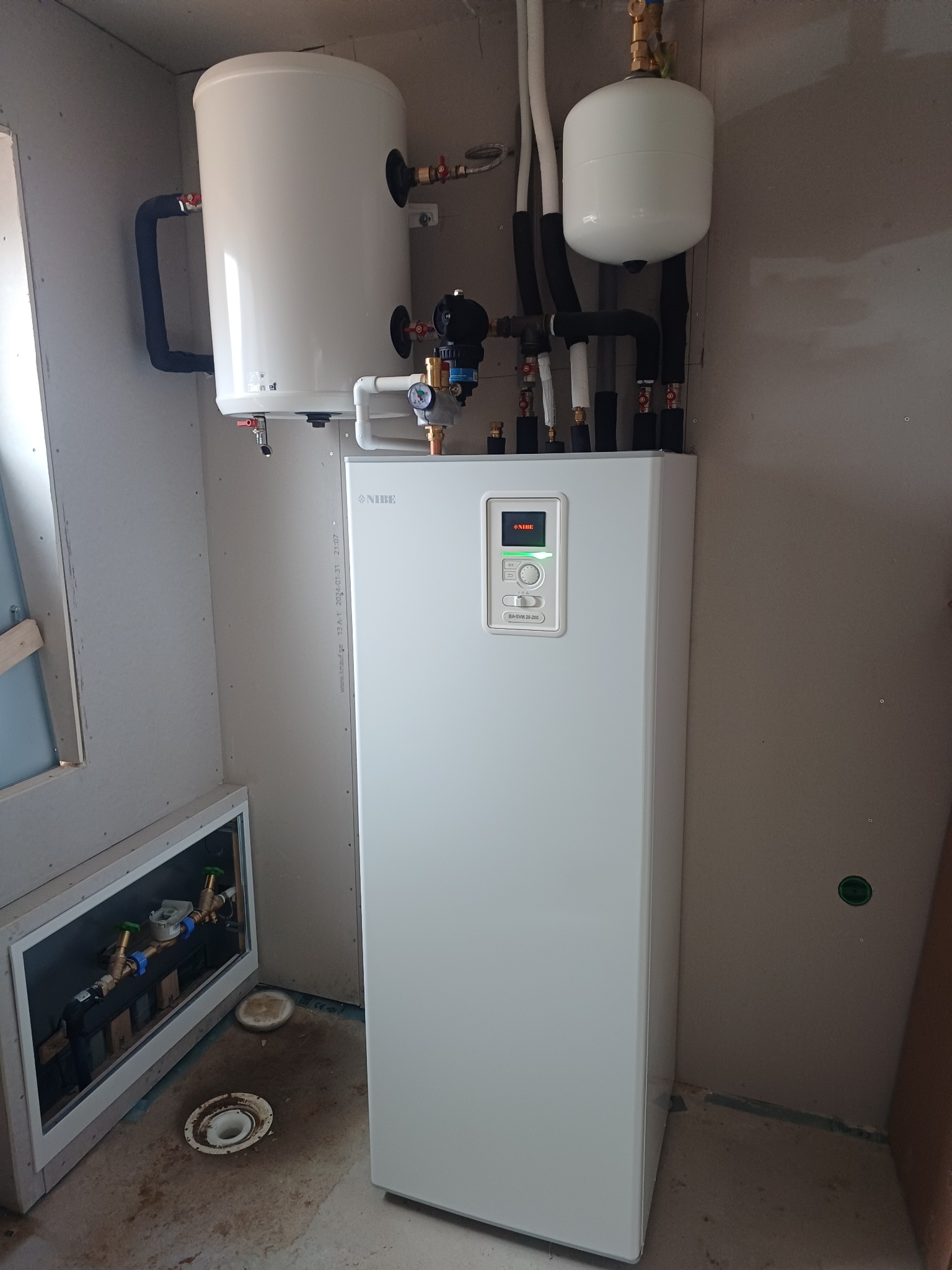 Biała pompa ciepła NIBE z zbiornikiem i osprzętem, zamontowana w pomieszczeniu z surowymi ścianami. Widoczne rury i zawory, instalacja w trakcie montażu.