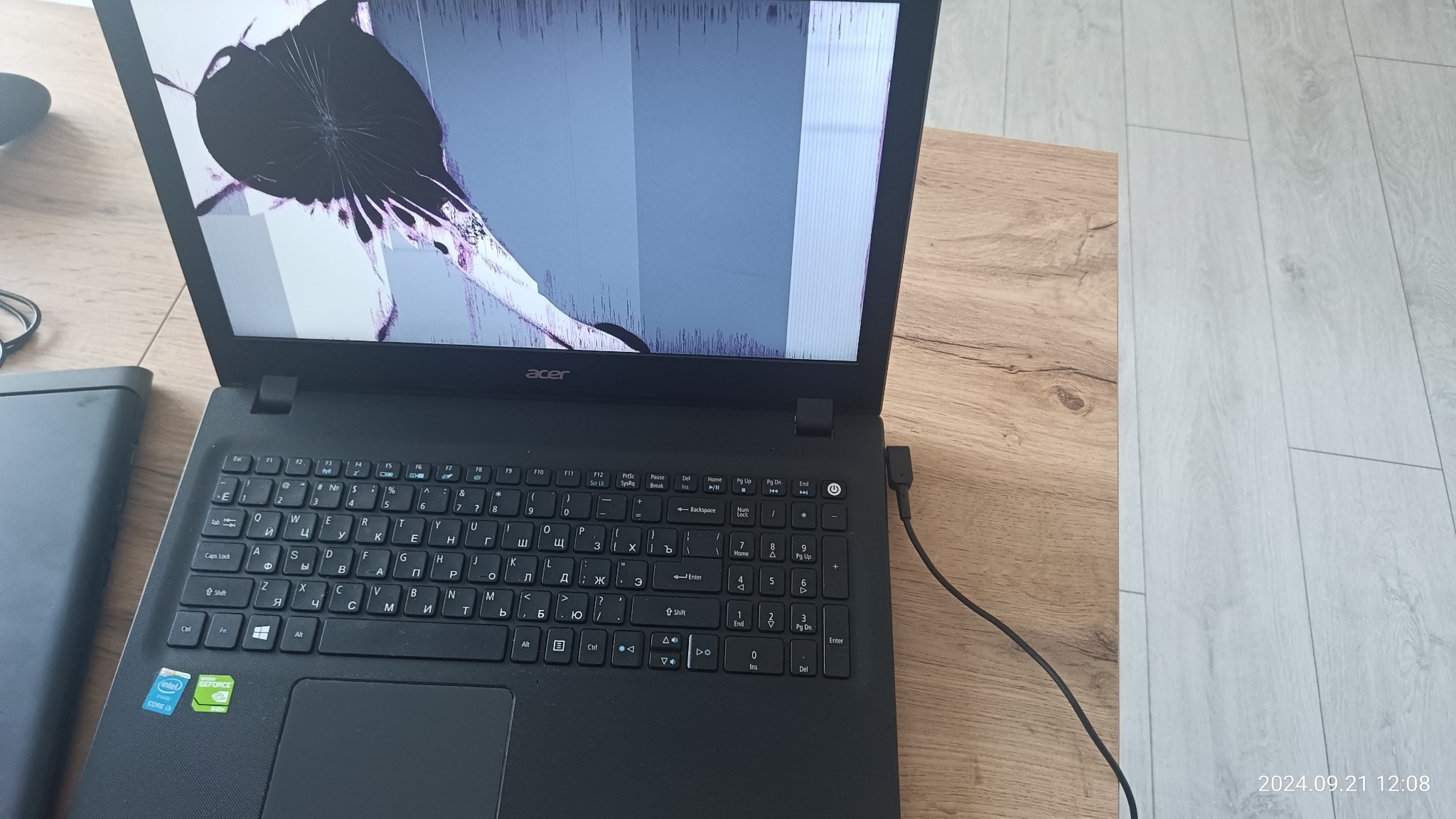 Czarny laptop Acer z wyraźnie pękniętym ekranem, widoczne uszkodzenia w kształcie pajęczyny, obok leży czarny dysk zewnętrzny, całość na drewnianym biurku.