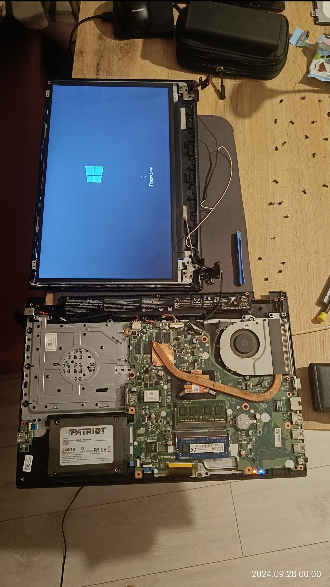 Otwarty laptop z widoczną płytą główną, procesorem z chłodzeniem, dyskiem SSD Patriot, odłączonym ekranem wyświetlającym logo Windows i rozsypanymi śrubkami na drewnianym biurku.