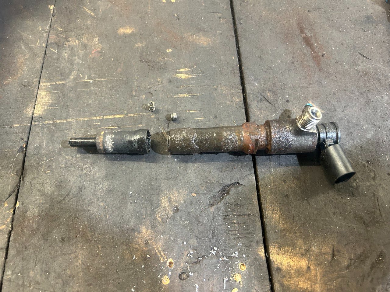 Rozłożony na części, brudny wtryskiwacz paliwa leżący na warsztatowym stole, widoczne sprężynki i ślady oleju.