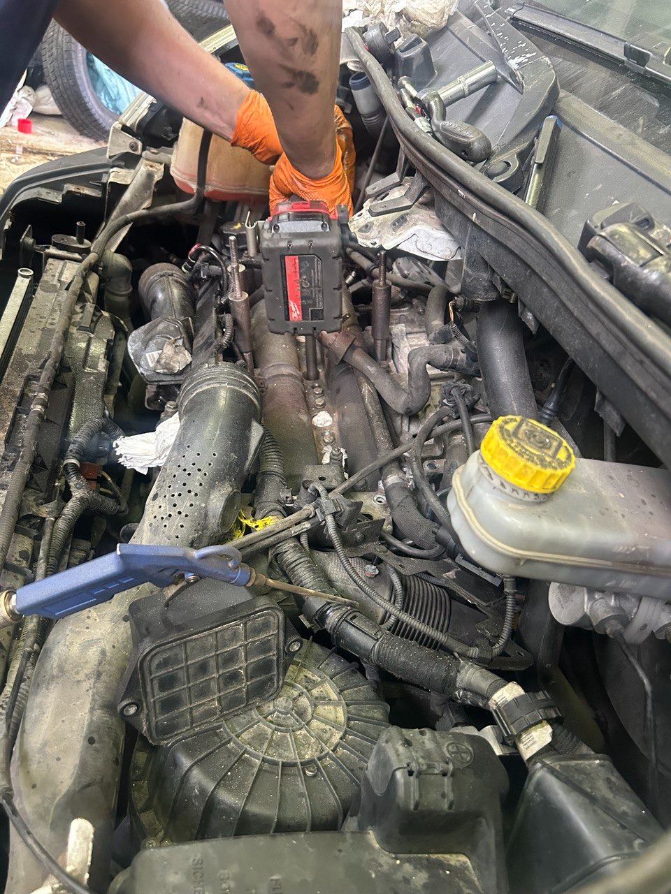 Mechanik w pomarańczowych rękawicach naprawia silnik samochodu używając narzędzia akumulatorowego Milwaukee, widoczne zdemontowane elementy i przewody pod maską.